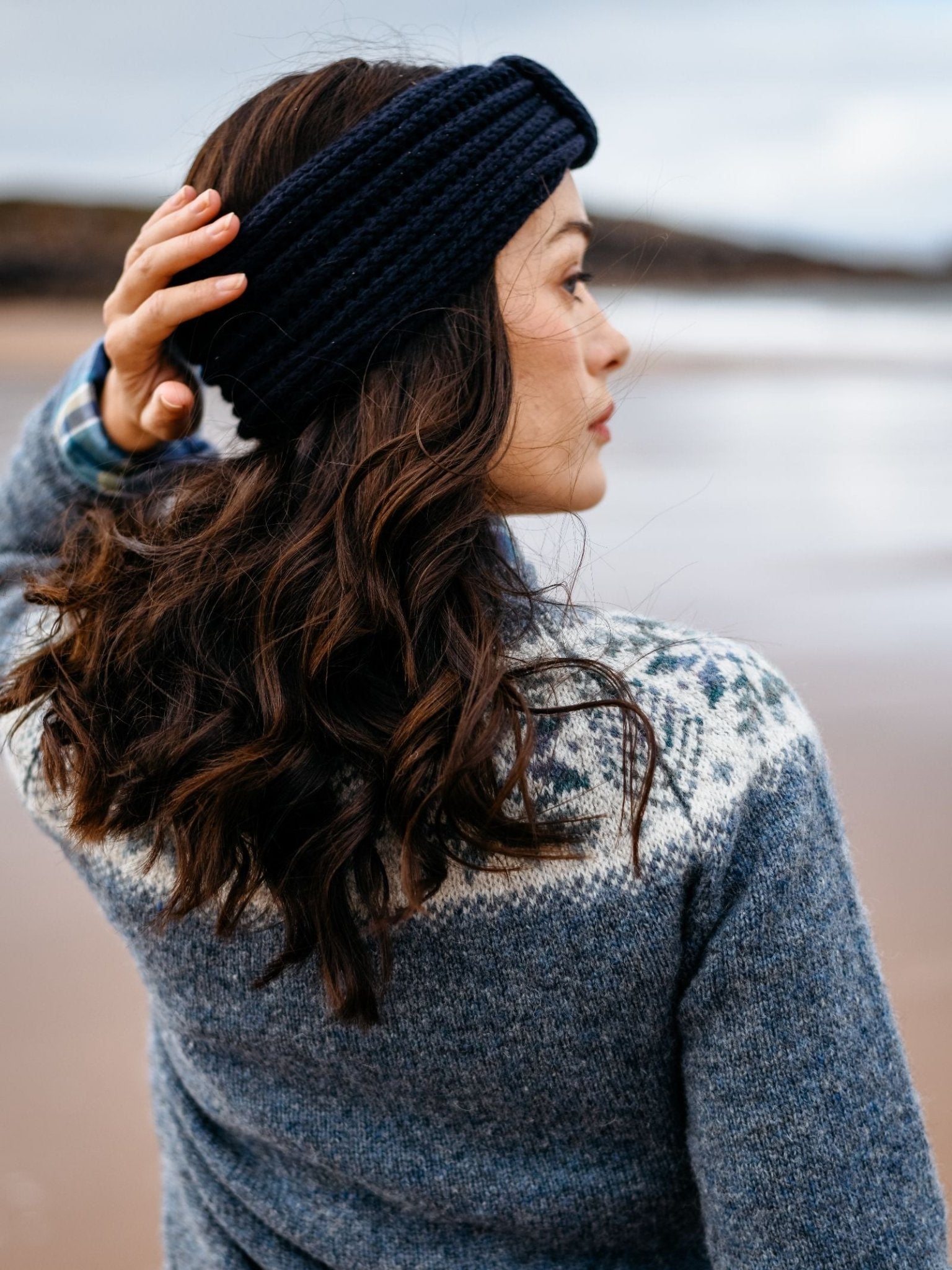 A woman with wavy brown hair wears a Campbell's of Beauly Cashmere Ear Warmer and a blue patterned sweater. She stands on the beach, facing away, touching her winter accessory as blurred sand and water fill the background.