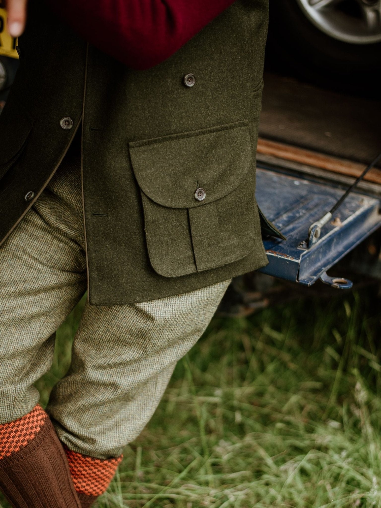 A person in a Campbell's of Beauly Luxury Field Nehru Gilet stands on grass by a vehicle’s open tailgate. The Loden green wool gilet features large pockets; light plaid trousers and rust socks complete the look. Upper body is partially out of frame.