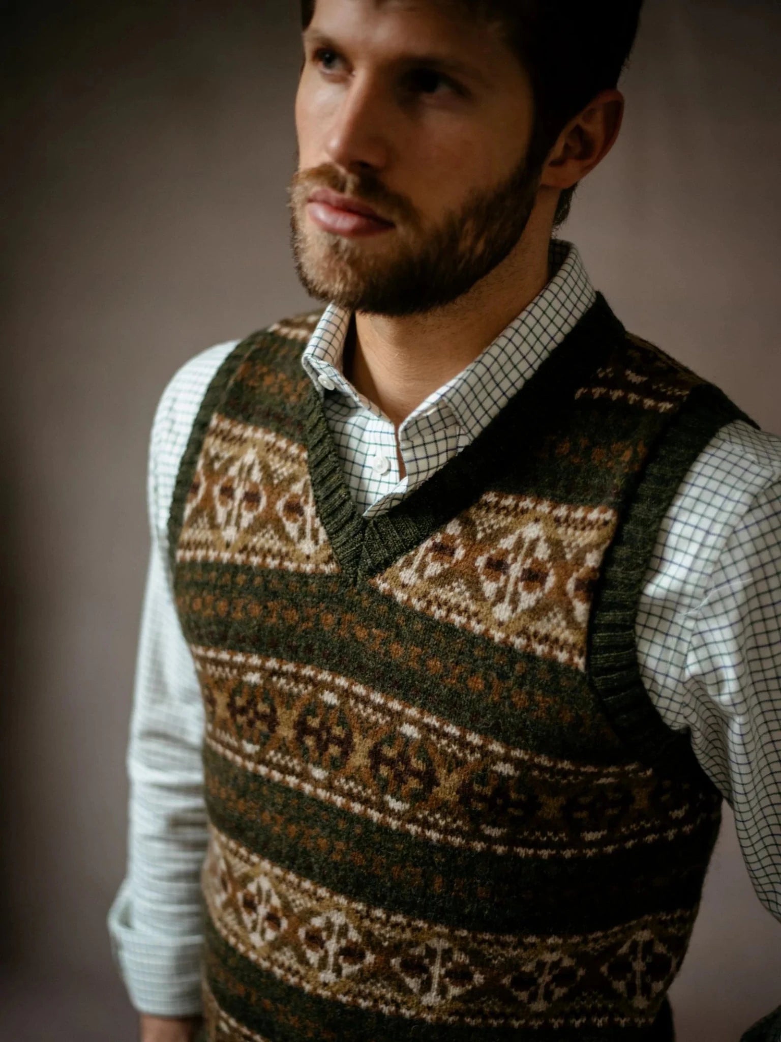 A man wearing the Campbell’s of Beauly Fairisle Slipover, crafted from soft Shetland wool, over a checkered shirt stands against a neutral background, gazing slightly away from the camera with a thoughtful expression.