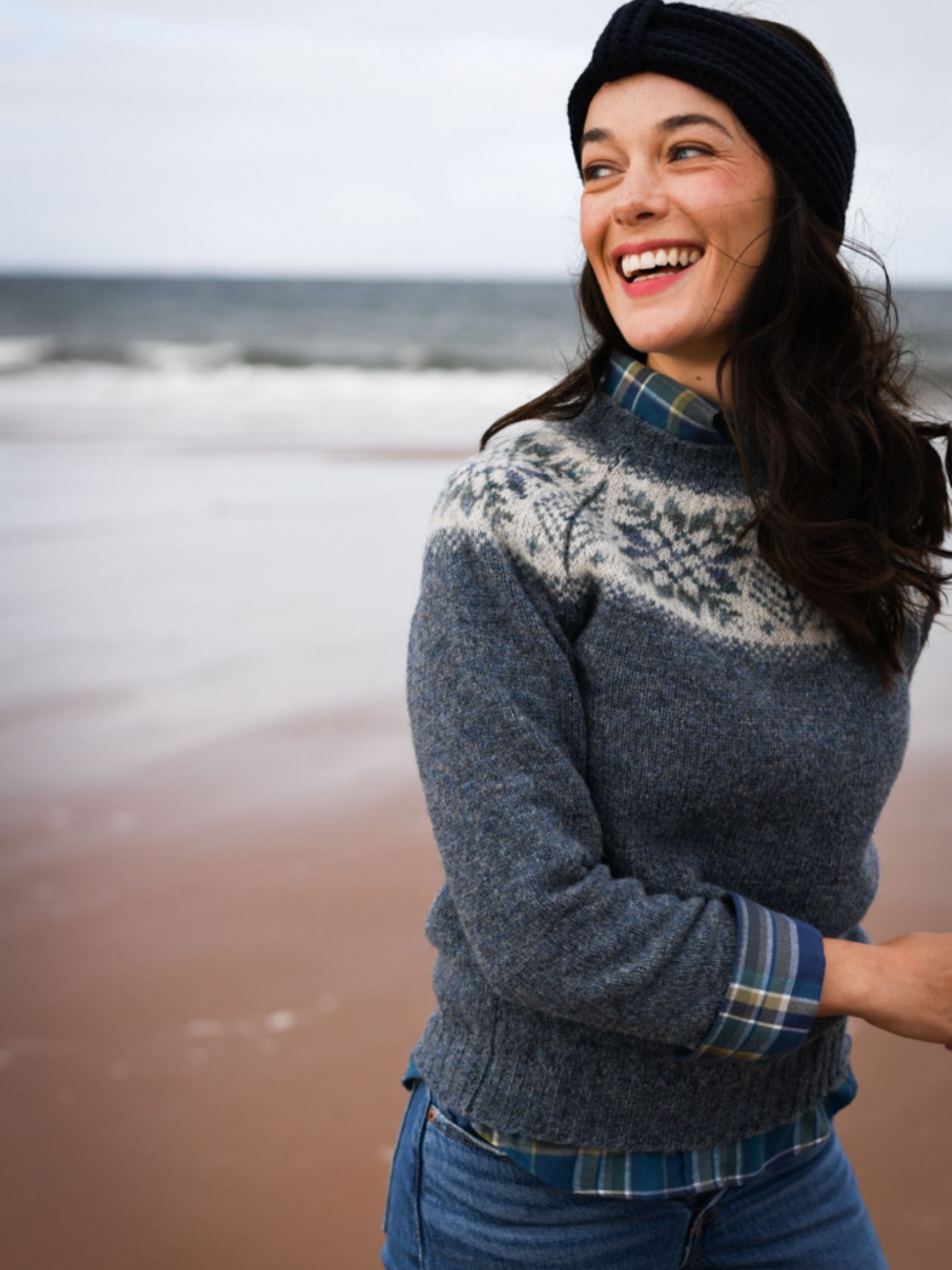 A woman wearing a Campbell's of Beauly Cashmere Ear Warmer, blue patterned sweater, and plaid shirt smiles on a sandy beach with the ocean behind her.