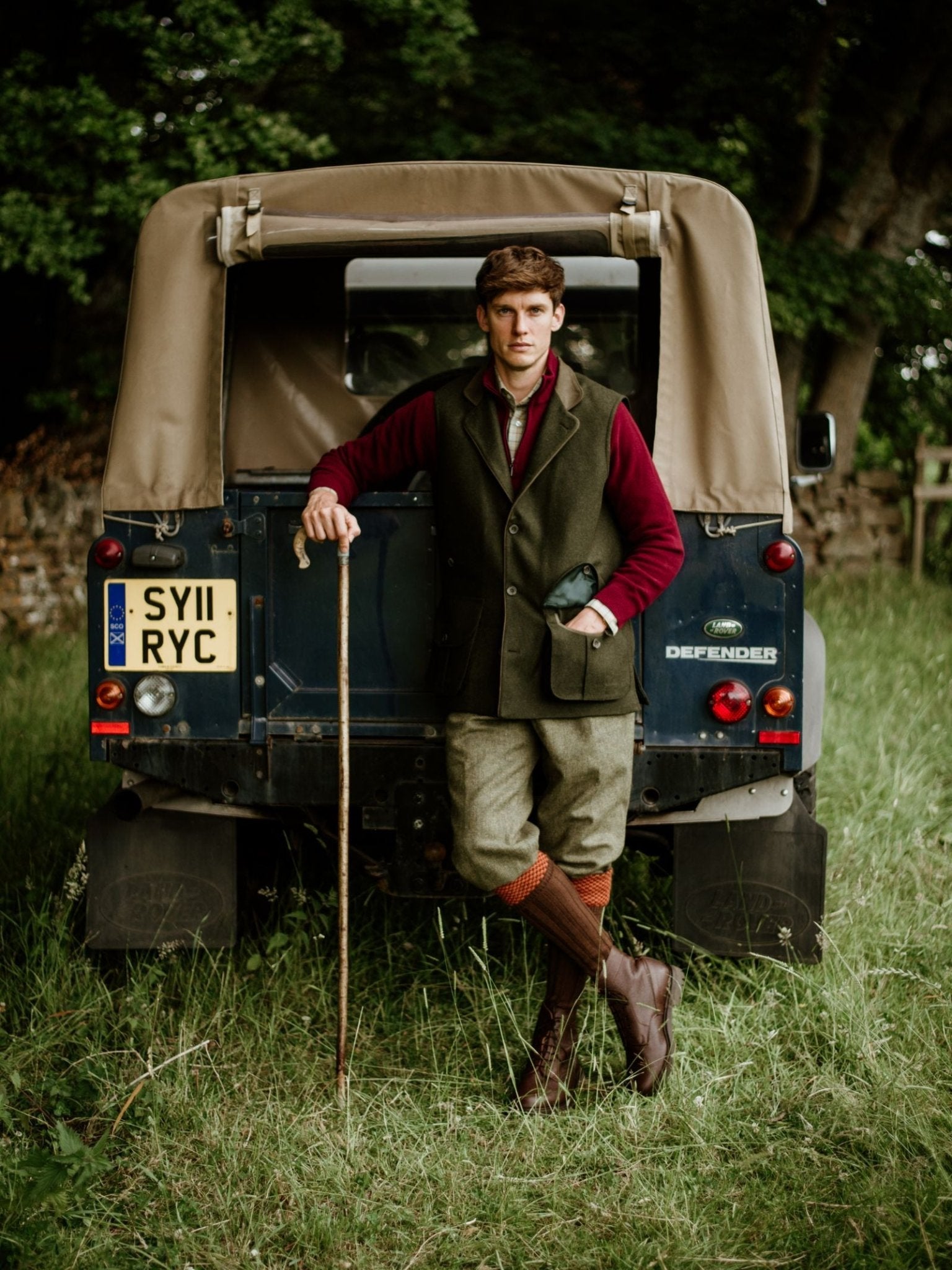 A man in a Campbell's of Beauly Luxury Field Nehru Gilet leans against a Land Rover Defender on grass, holding a walking stick with trees and stone walls behind him.