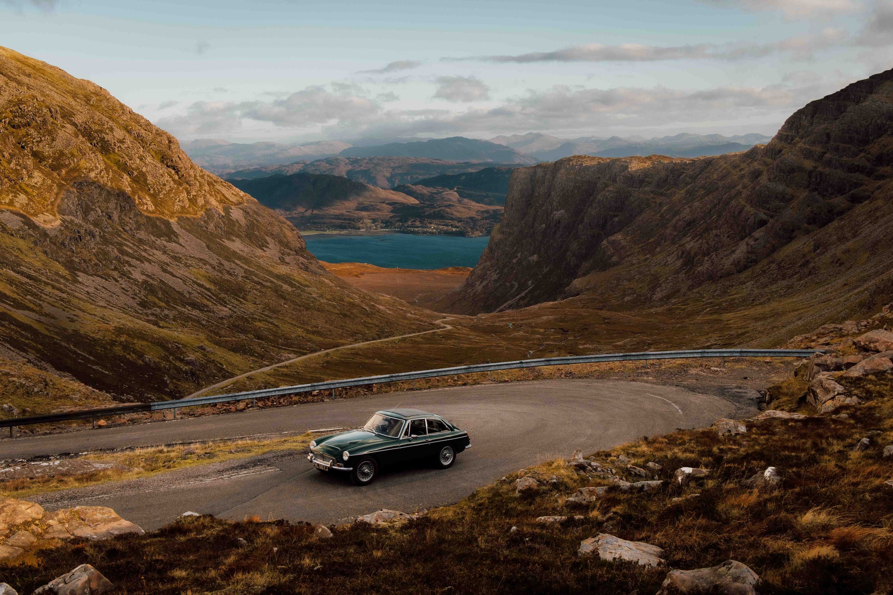 A classic dark green car drives on a winding mountain road, surrounded by rocky hills and valleys with a distant lake and mountains under a partly cloudy sky.
