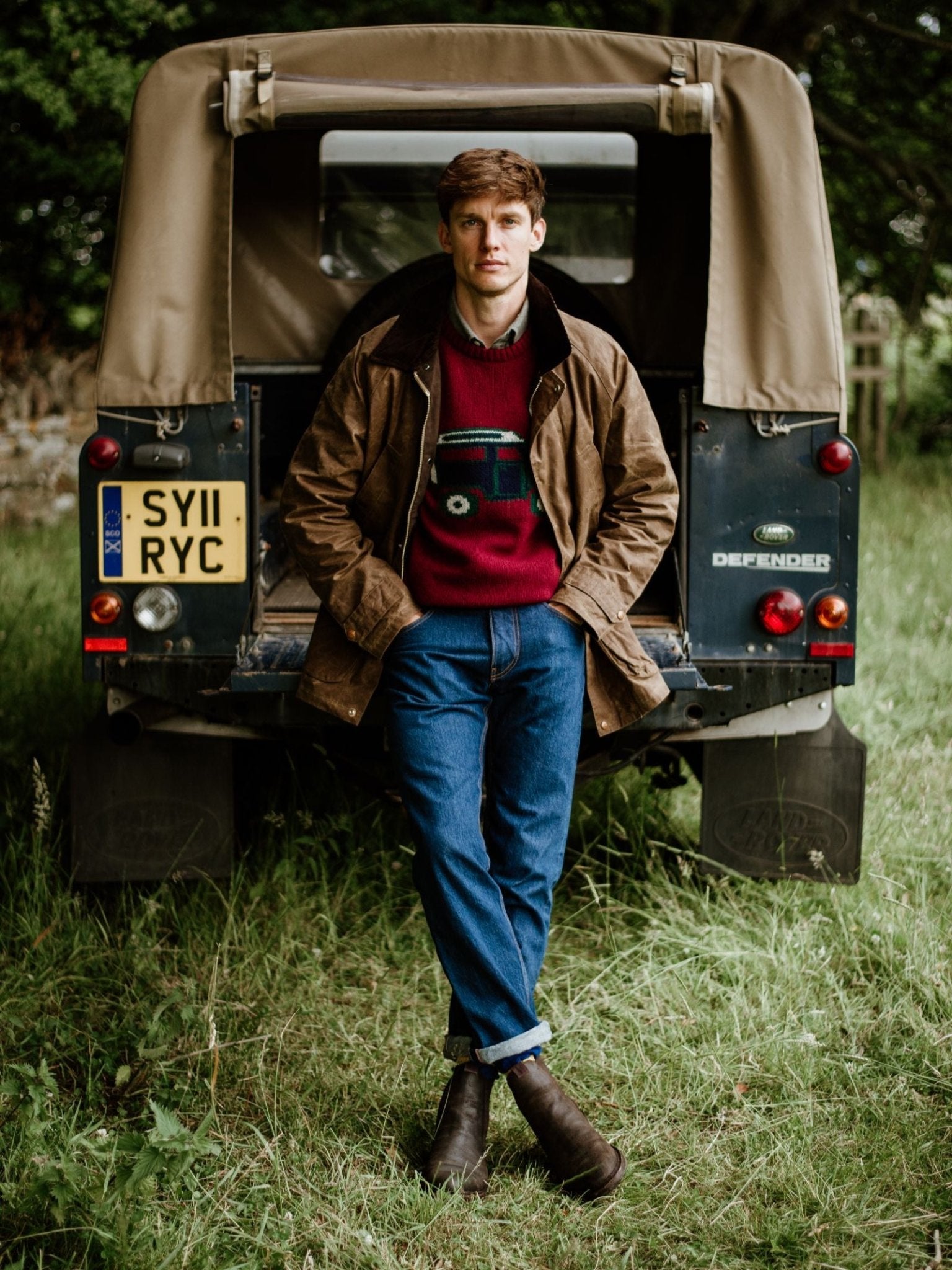 A man in a brown jacket, red sweater, blue jeans, and Campbell's of Beauly Lowlander Boots stands with arms crossed in front of a green Land Rover Defender with a tan roof, parked on grass, looking at the camera.