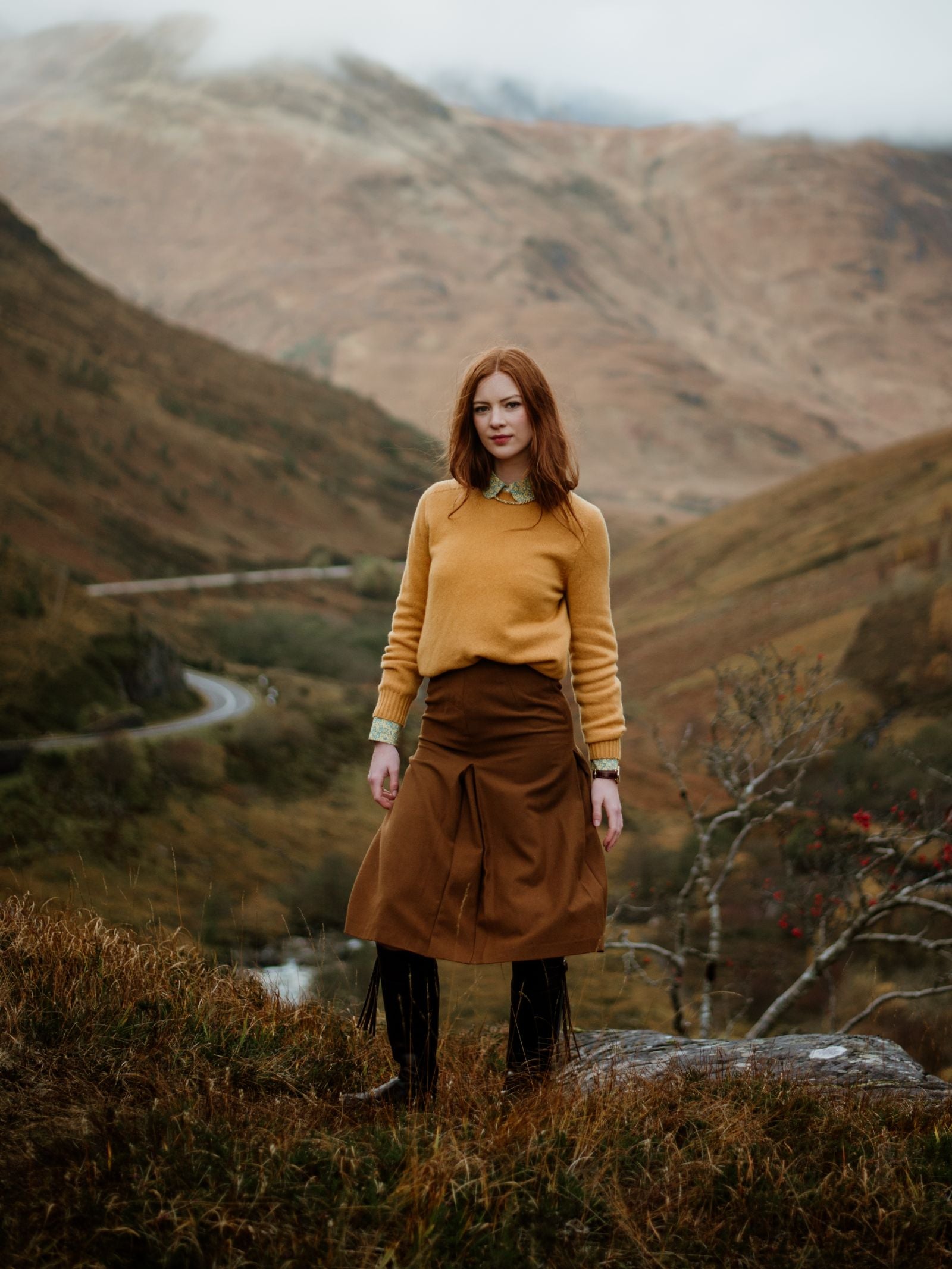 A red-haired woman stands outdoors on grass, wearing a mustard Auld Stock Geelong Lambswool Crew Neck Jumper by Campbell's of Beauly and a brown skirt. Rolling hills and a winding road are behind her beneath a cloudy sky.