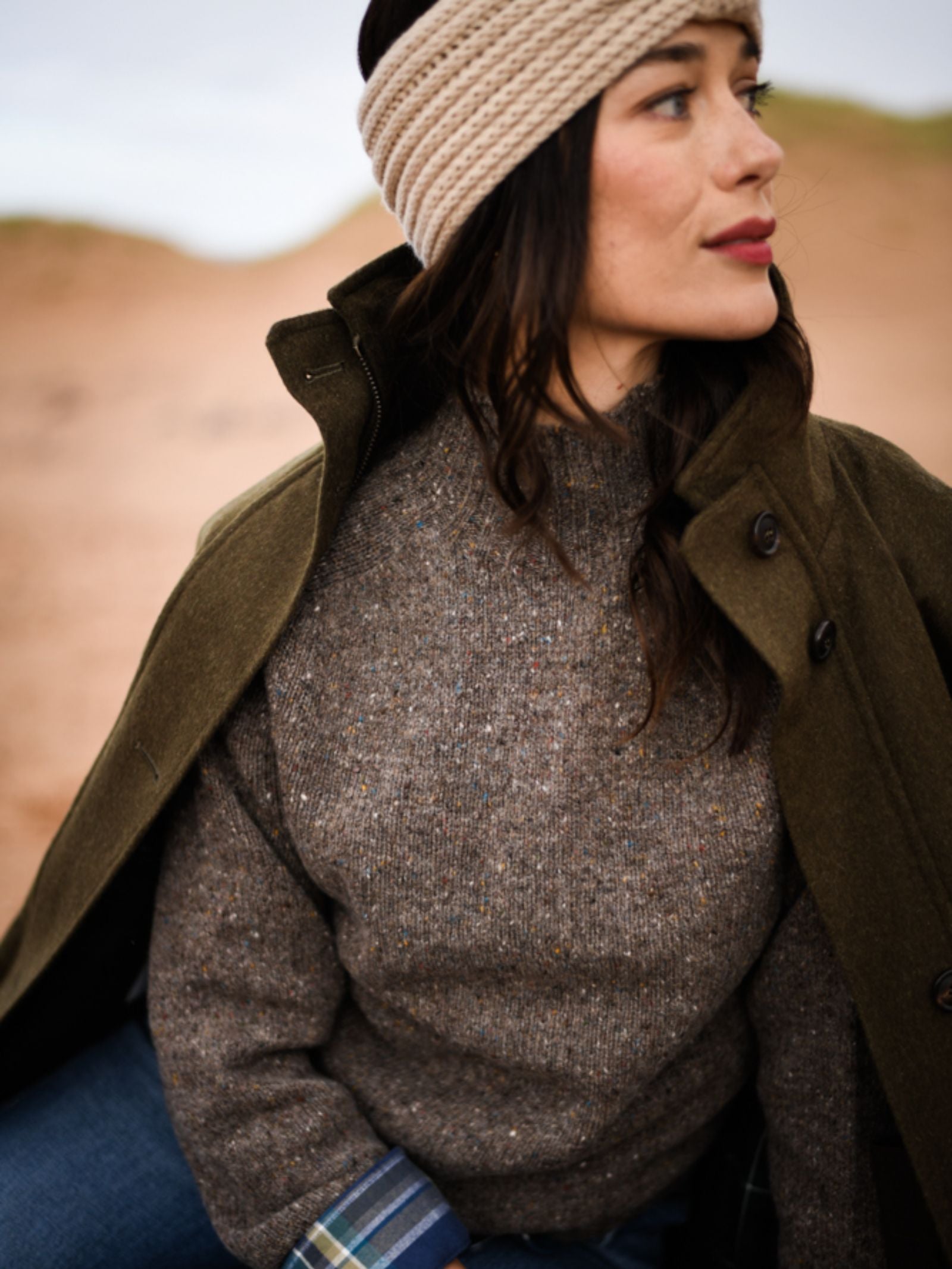 A woman in a beige knitted headband, blue jeans, olive green coat, and the Donegal Lambswool Funnel Neck Jumper by Campbell's of Beauly sits outdoors with a calm expression. The earthy-toned background is softly blurred.