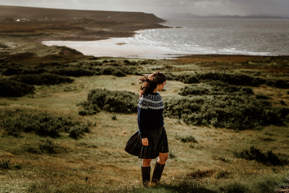 A woman in a knit sweater, plaid skirt, and boots stands on a grassy hill overlooking a rocky coastline and the sea, with waves and distant land visible under a cloudy sky.