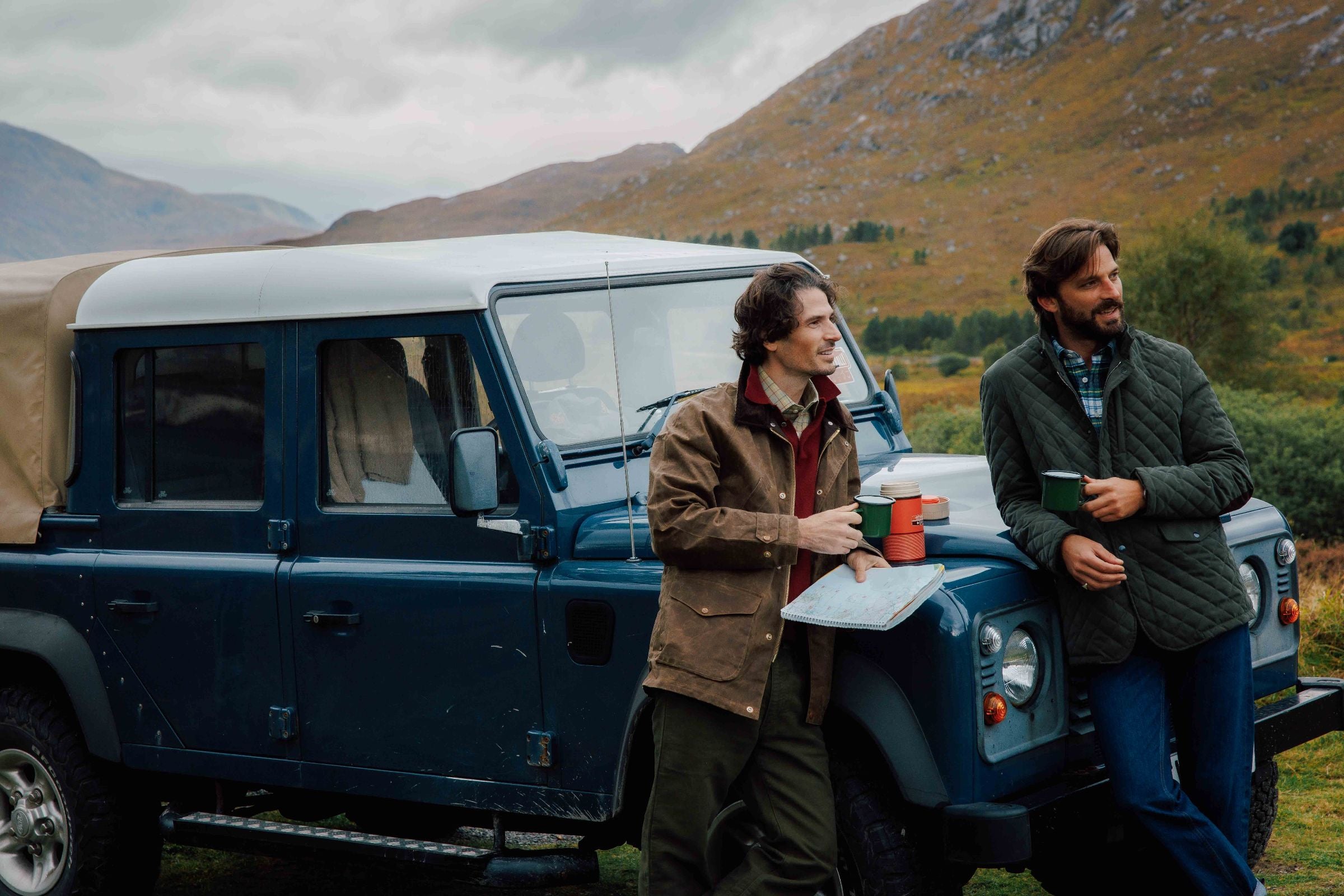 Two men stand by a blue off-road vehicle parked in a scenic, mountainous area. One holds a map and a red cup, while the other holds a green cup. Both appear relaxed and are dressed in casual outdoor clothing.