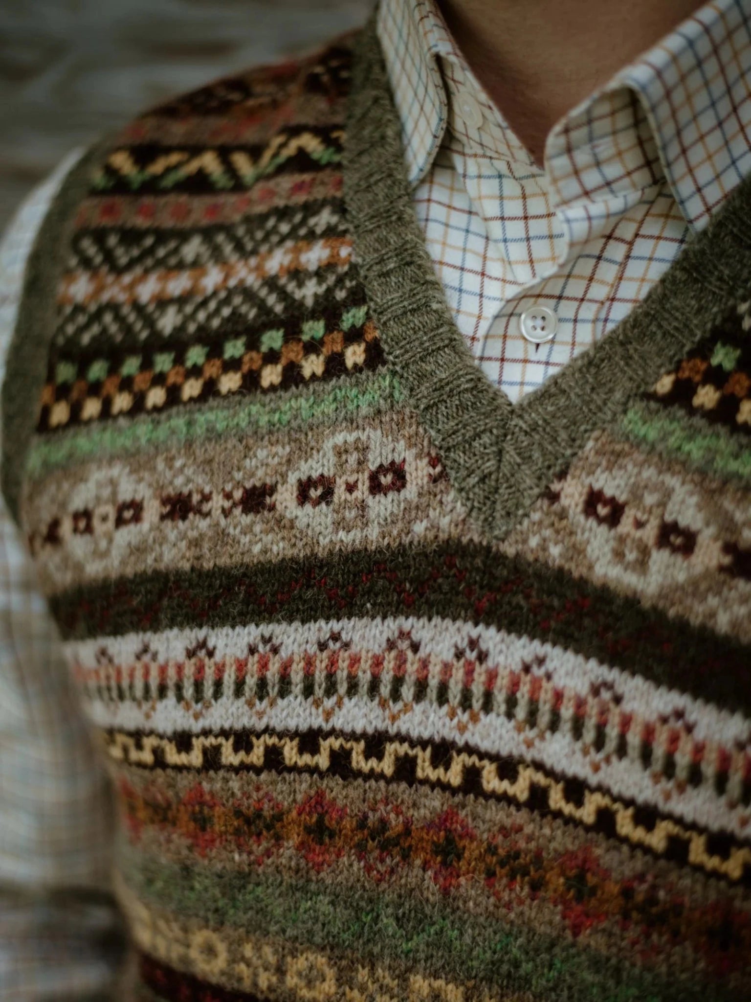 Close-up of a person wearing the Campbell’s of Beauly Fairisle Slipover in earth tones, crafted from soft Shetland wool and layered over a white plaid button-up shirt. Only the chest and part of the collar are visible, emphasizing this classic sleeveless layer.