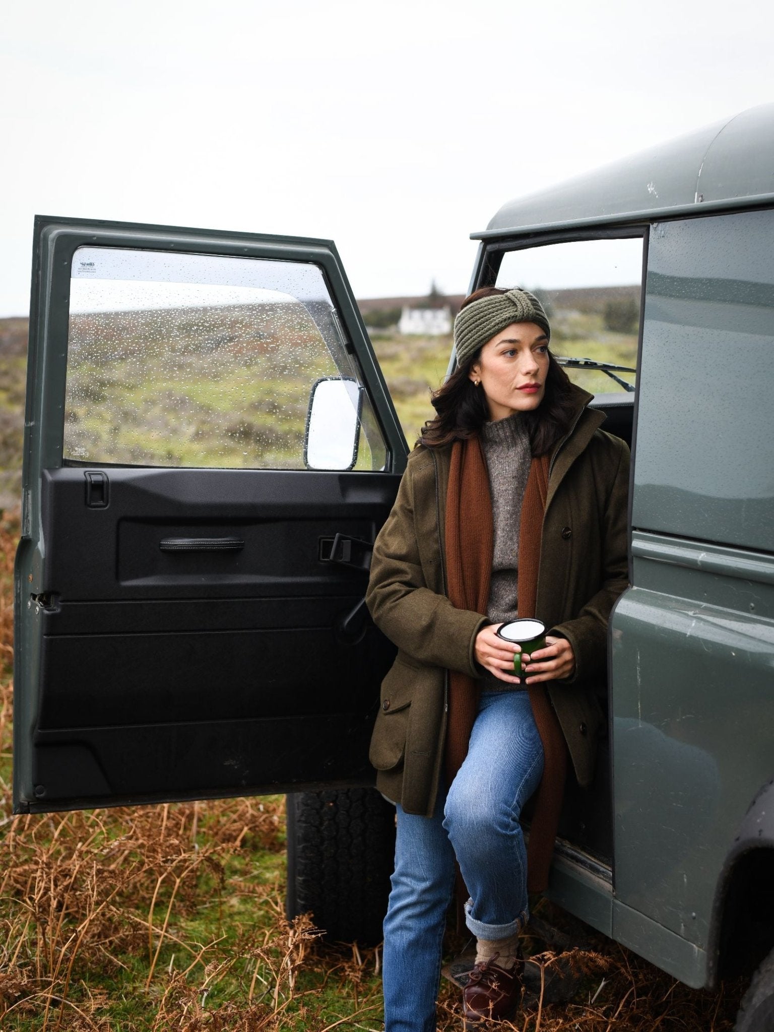 A woman wearing a Campbell's of Beauly Cashmere Ear Warmer, green jacket, and brown scarf leans against an open car door holding a cup in a grassy rural area with a house in the background.