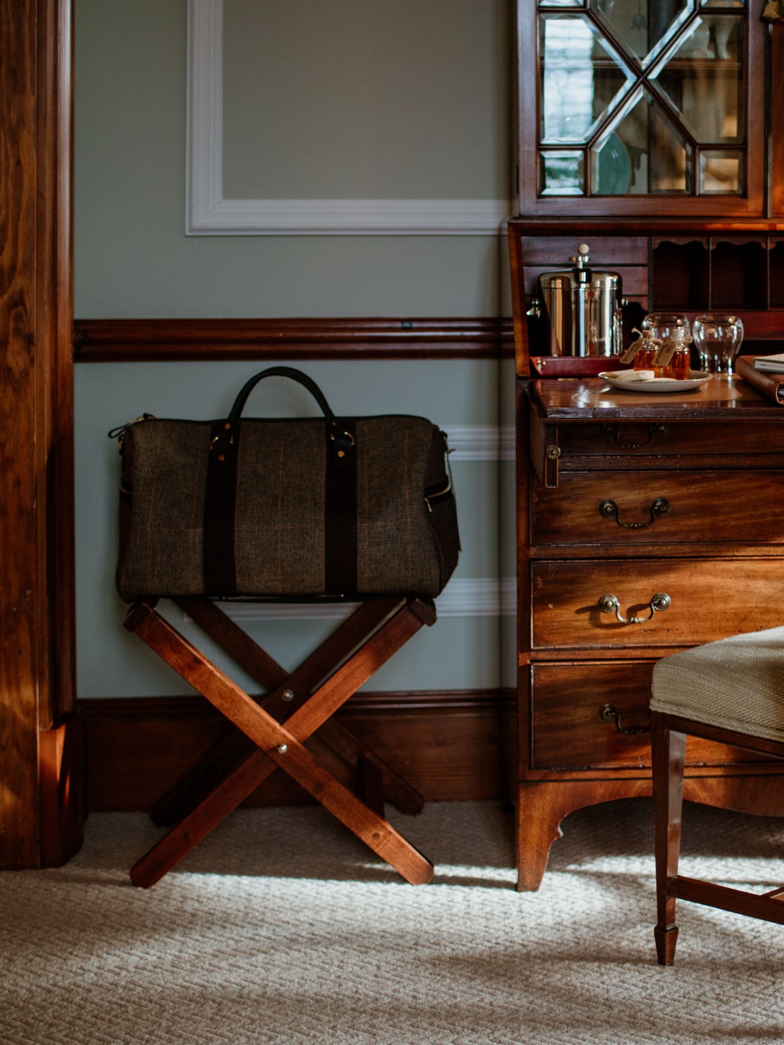 A Campbell's of Beauly Hand Luggage Holdall in brown vegetable-tanned leather rests on a wooden luggage rack in a warmly lit room with classic wood furniture and a chest of drawers topped with glasses and an ice bucket.