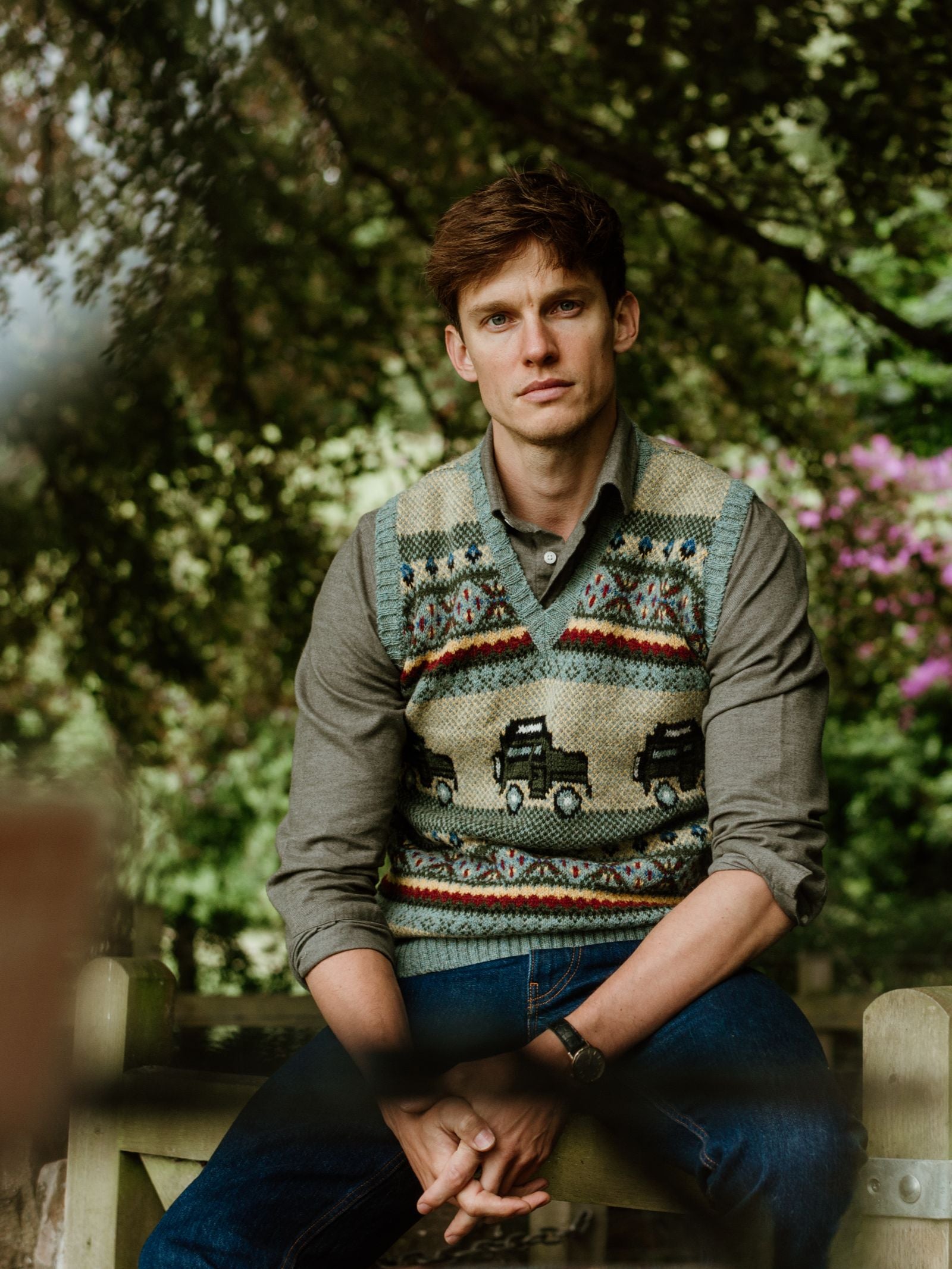 A young countryside enthusiast with short brown hair sits outdoors on a wooden bench, wearing a gray shirt and the Campbell's of Beauly Land Rover Defender Fairisle Slipover, surrounded by trees and greenery as he looks calmly at the camera.