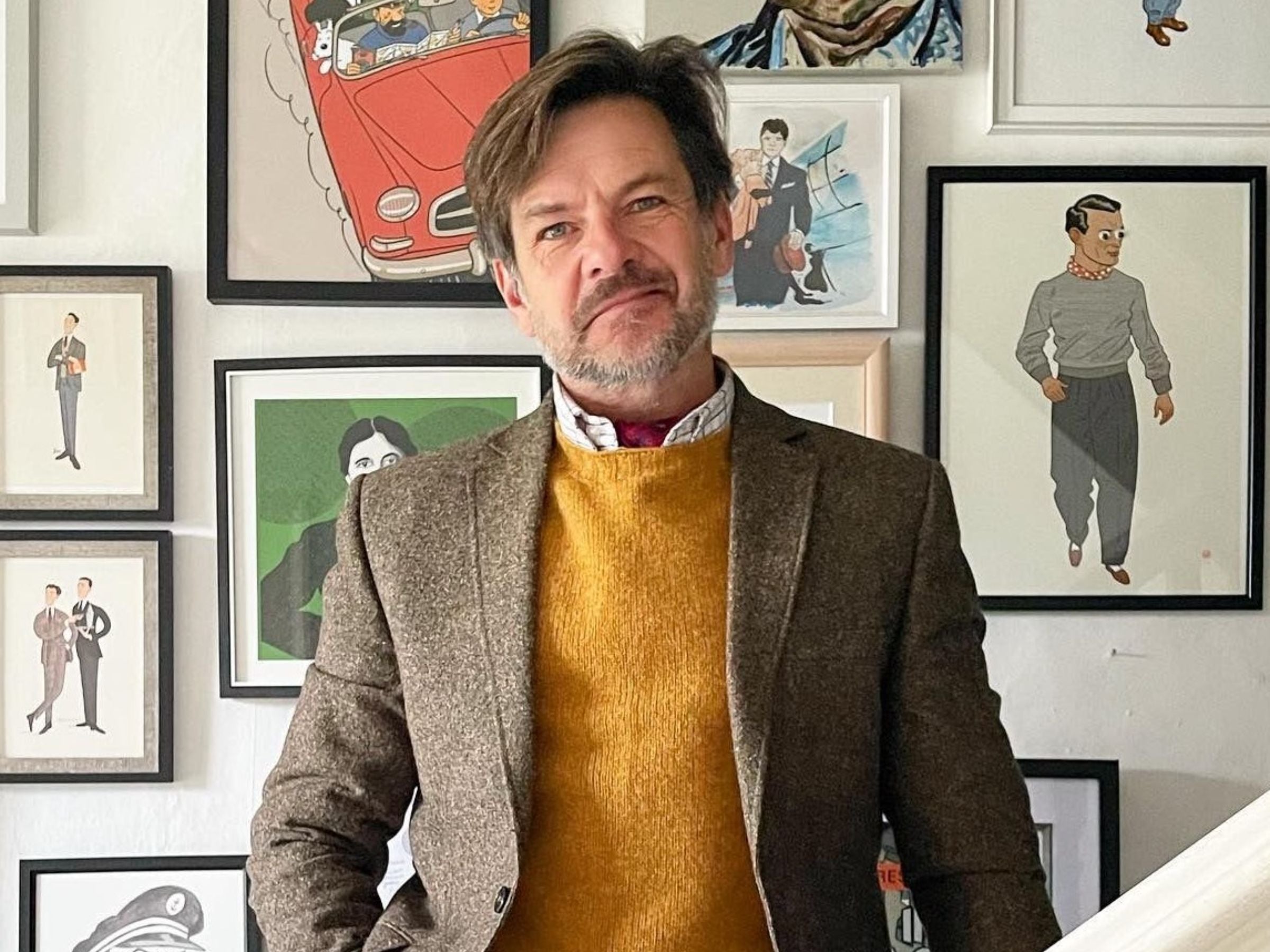 A man with short brown hair and a beard, wearing a brown blazer and mustard yellow sweater, stands indoors in front of a wall decorated with various framed illustrations and artwork.