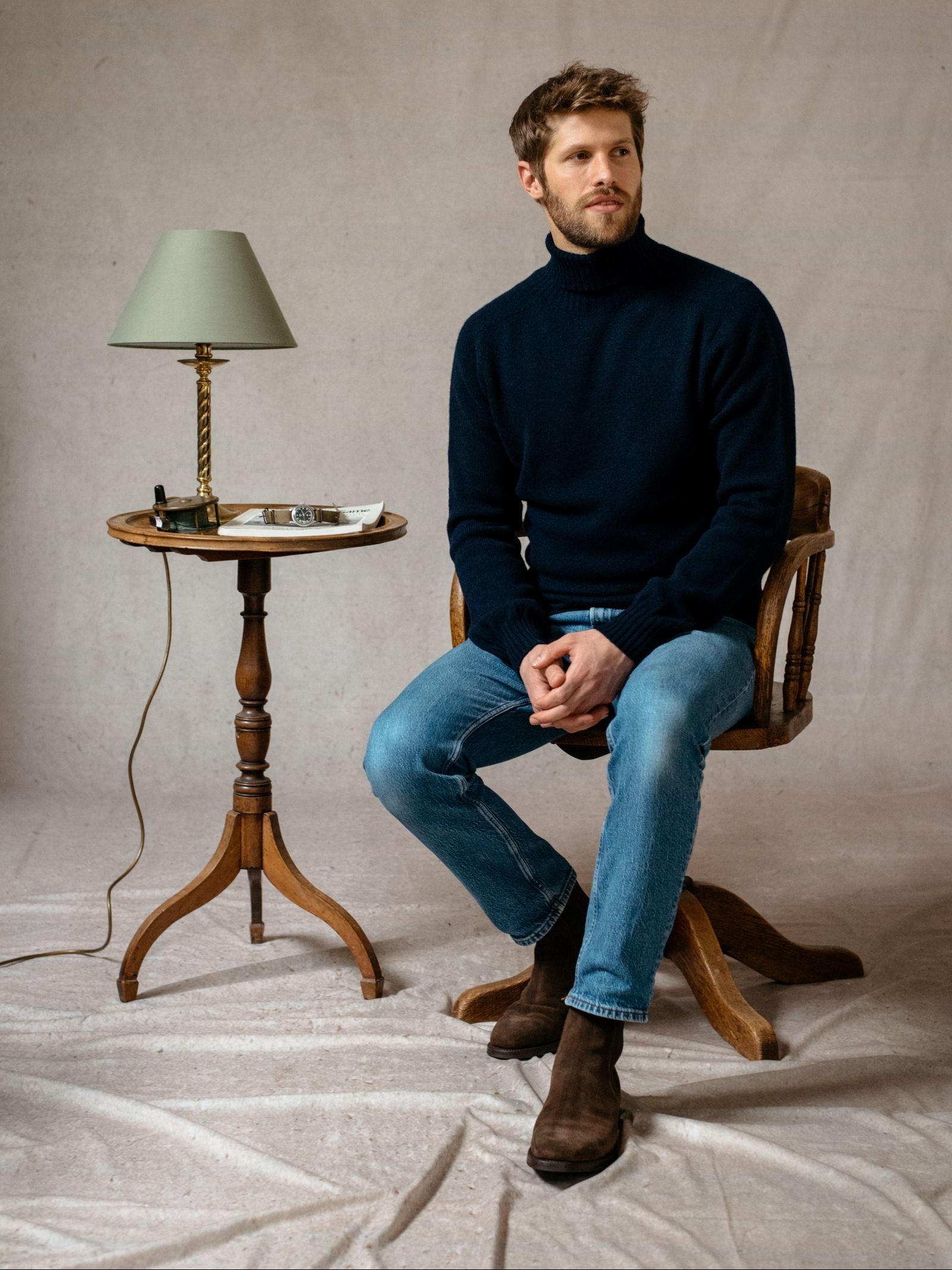 A man in Campbell’s of Beauly Lambswool Roll Neck and blue jeans sits on a wooden chair by a table with a green lamp, notebook, and pen against a neutral backdrop—the ideal layering piece for any occasion.
