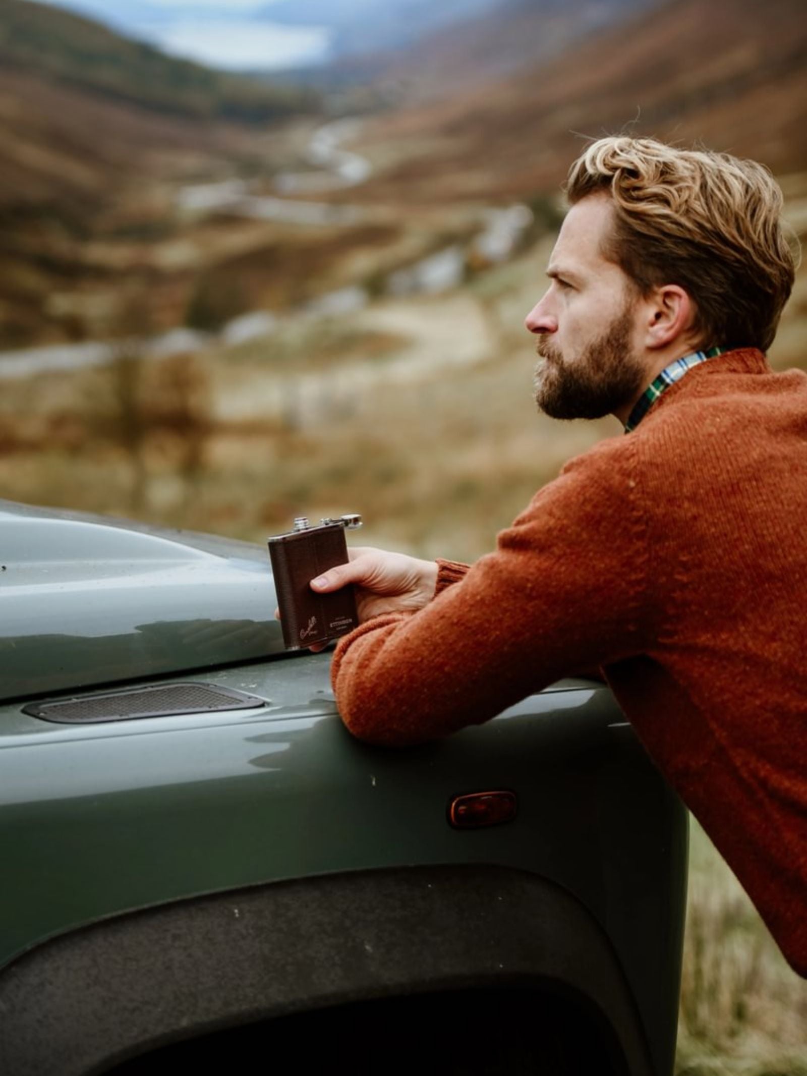 A man with blond hair and a beard, in a rust-colored sweater, leans on a green vehicle holding the 6oz Chocolate Hip Flask by Campbell's of Beauly x Ettinger, gazing thoughtfully into a scenic, hilly landscape reflecting British craftsmanship.