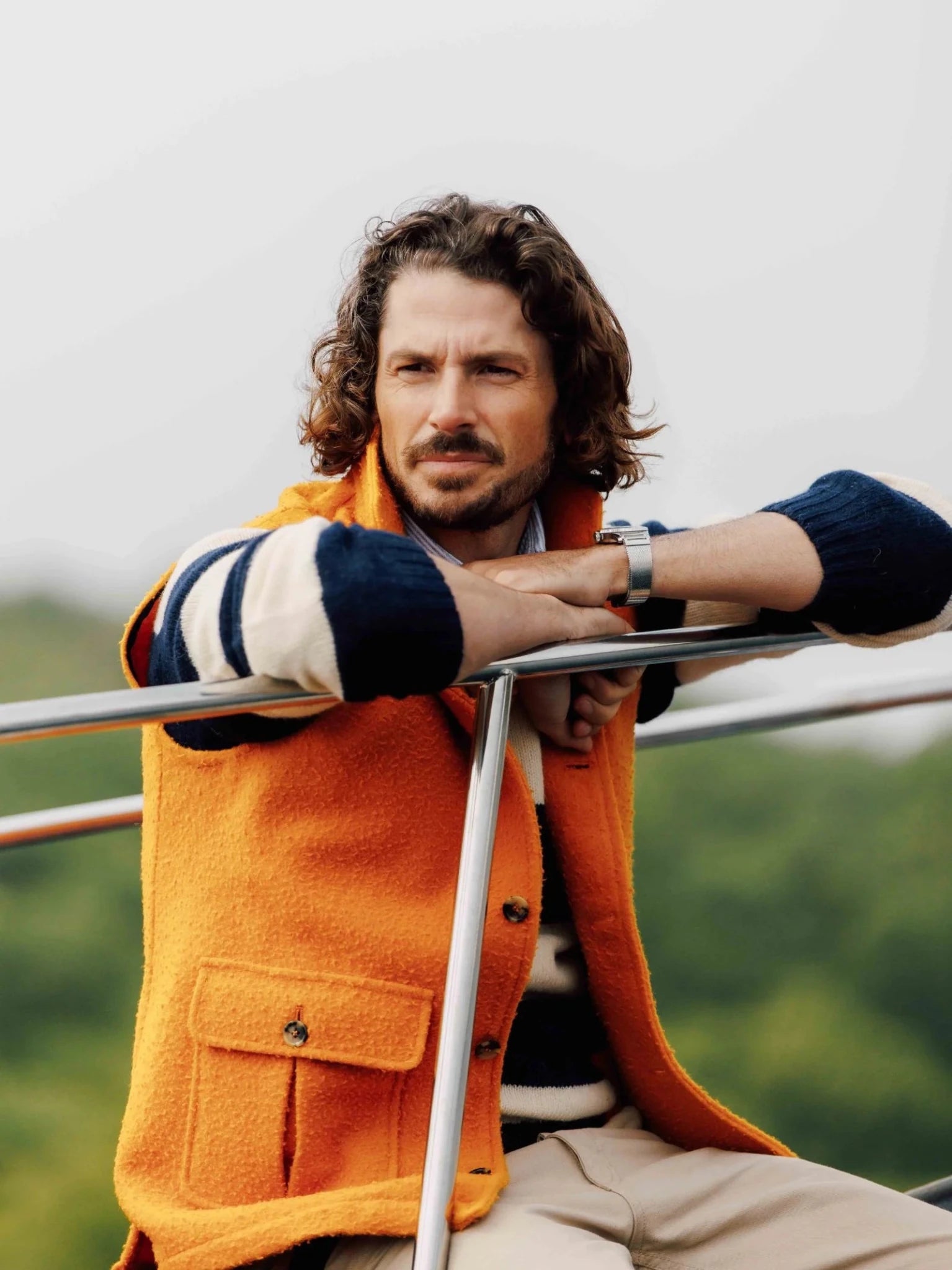 A man with wavy brown hair and a beard leans on a metal railing outdoors, wearing the Campbell's of Beauly Auld Stock Casentino Fleece Gilet in bright orange over a navy and white striped sweater, with green trees and an overcast sky behind him.