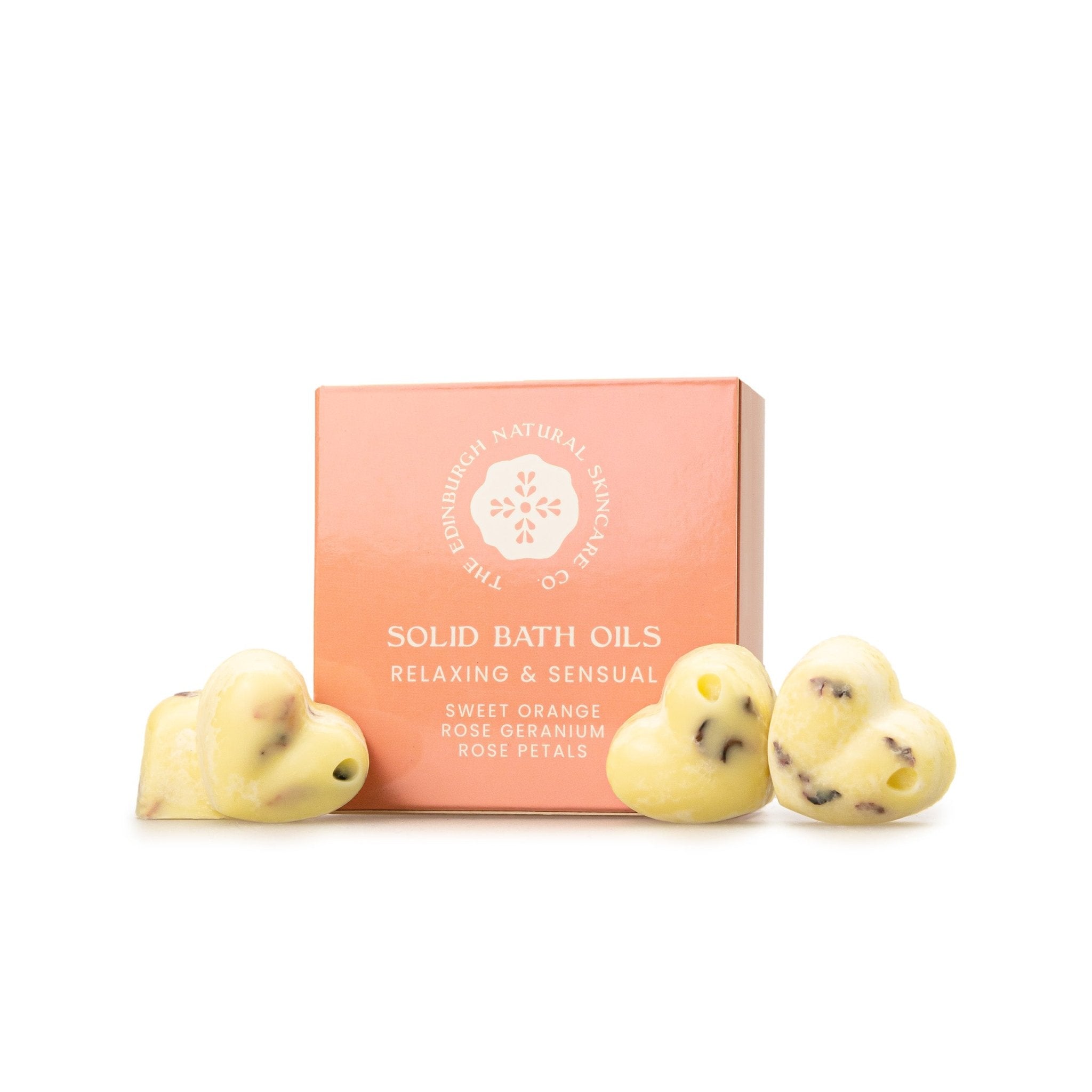 A pink box labeled Sweet Orange & Geranium Solid Bath Oils by The Edinburgh Natural Skincare Company is shown with three yellow, heart-shaped natural bath oils infused with visible flower petals in front of it.
