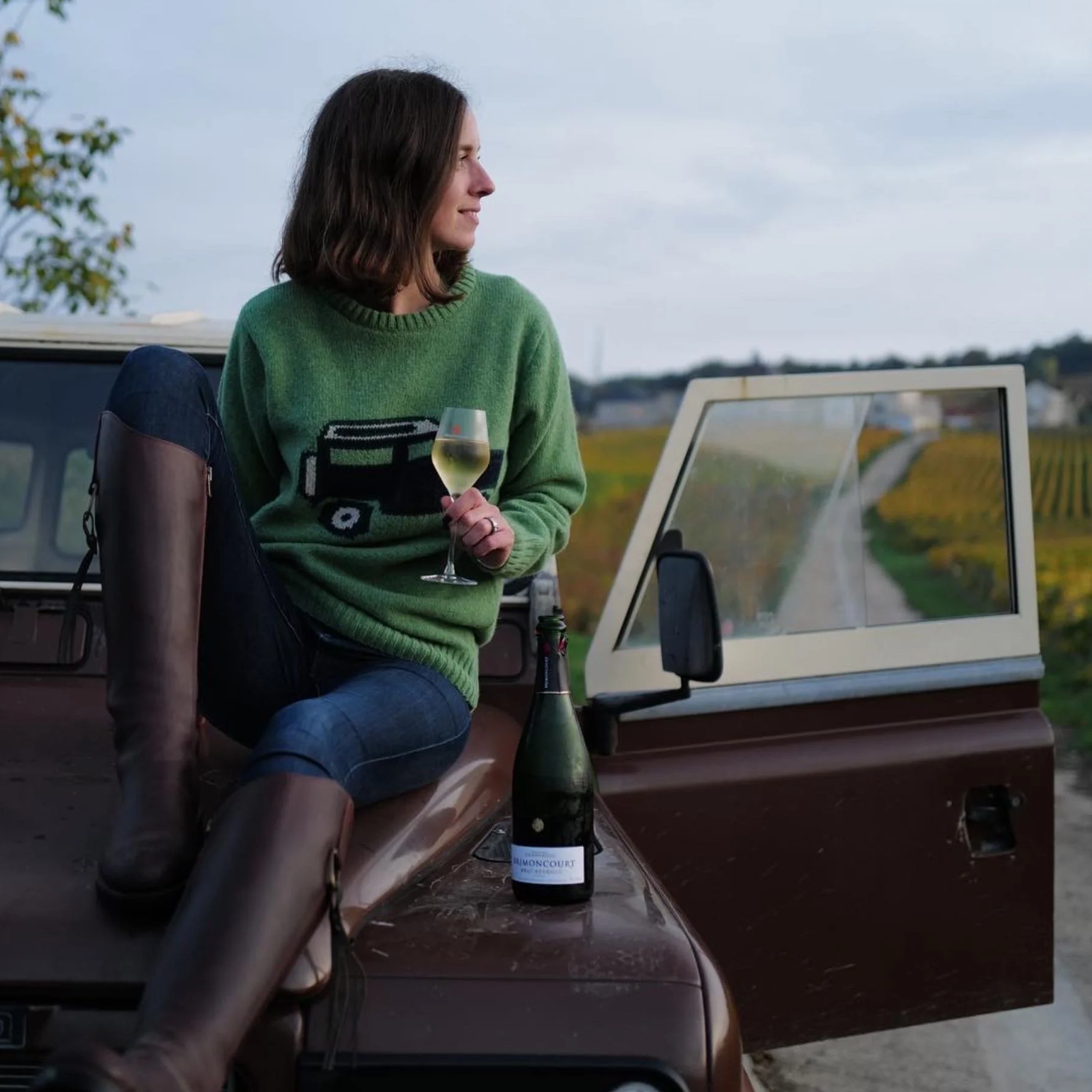 A woman in a green sweater and tall boots sits on the hood of a brown car, holding a glass of white wine. An open car door reveals a rural landscape with vineyards and a dirt road in the background.