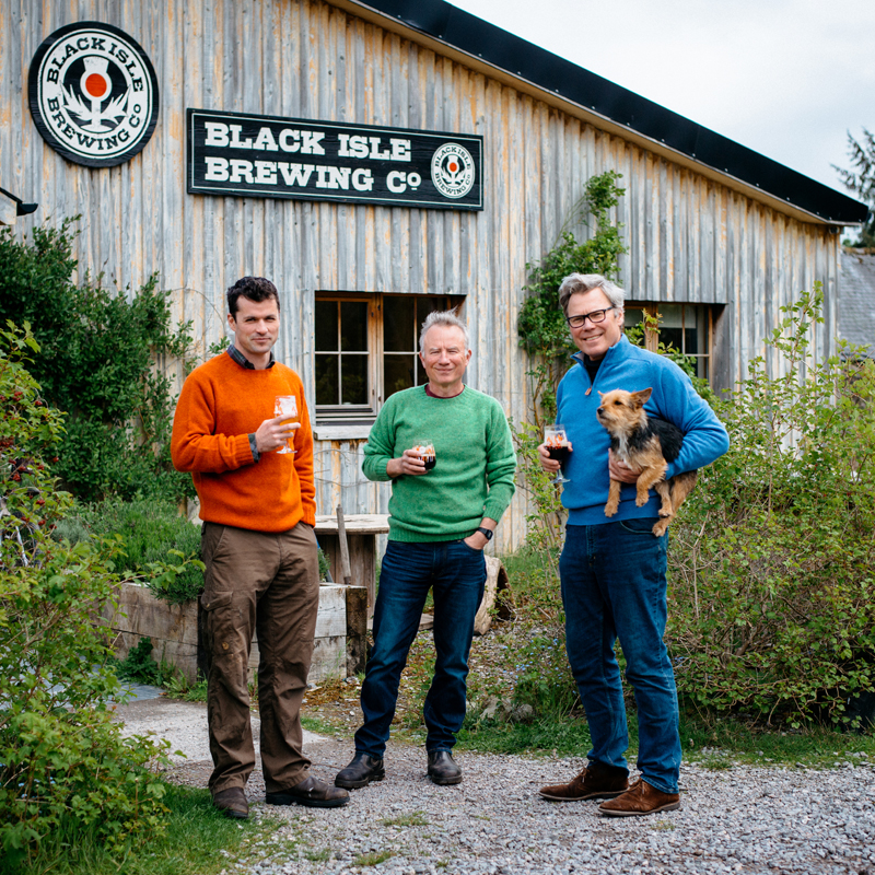 Three men stand outside the Black Isle Brewing Co. building, each holding a drink. One man holds a small dog. They are dressed in colorful sweaters, and greenery surrounds them. The building has wood paneling and signs.
