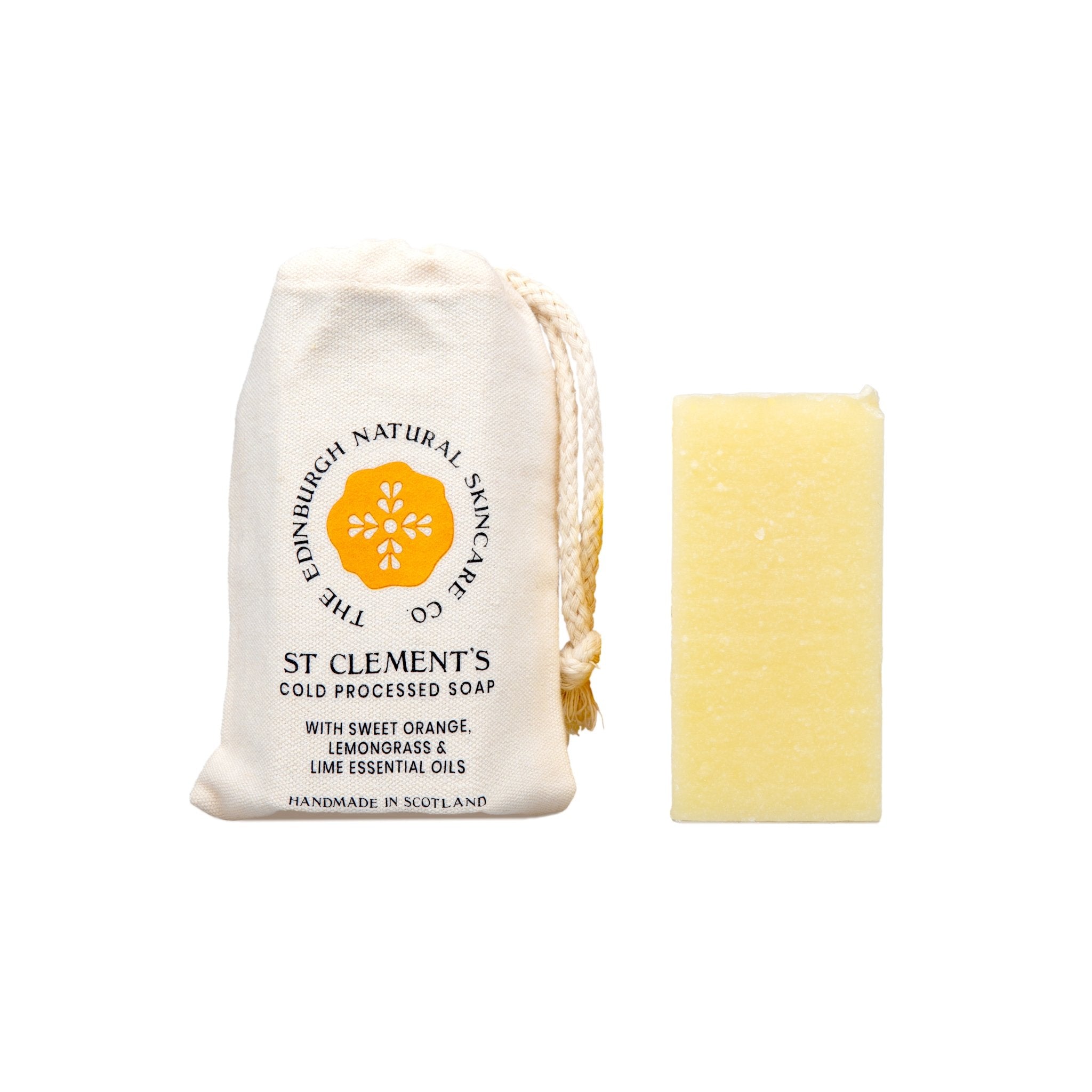 A rectangular yellow bar of St Clements Cleansing Soap by The Edinburgh Natural Skincare Company sits beside a cream drawstring bag, featuring a citrus-inspired design that highlights its natural orange, lemongrass, and lime oils.
