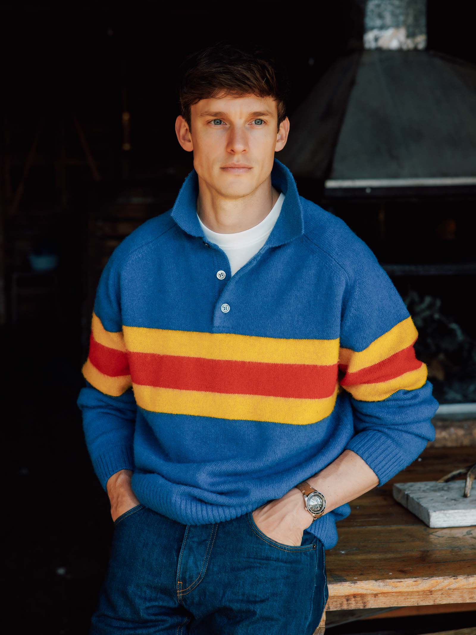 A young man with short brown hair wears the Campbell's of Beauly Brushed Shetland Stripe Rugby in blue, paired with blue jeans and a watch. He stands indoors with one hand in his pocket, looking confidently at the camera.