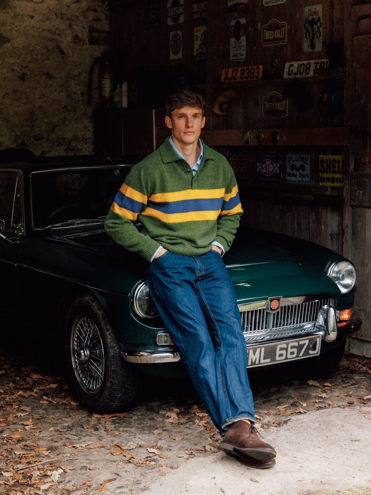 A young man in Campbell’s of Beauly Brushed Shetland Stripe Rugby, blue jeans, and brown shoes leans against a classic dark green car parked in a rustic garage with vintage signs on the wooden wall behind him.