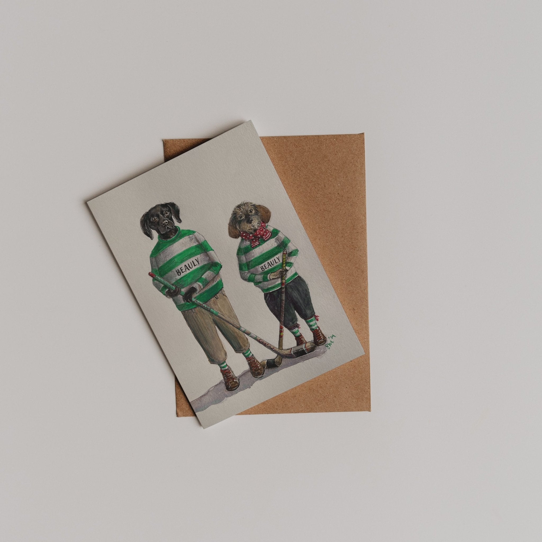 The Beauly Shinty Players Greeting Card by Campbell’s of Beauly features two dogs in green and white striped sweaters and pants, side by side, with a brown envelope. Inspired by the brand’s signature style.