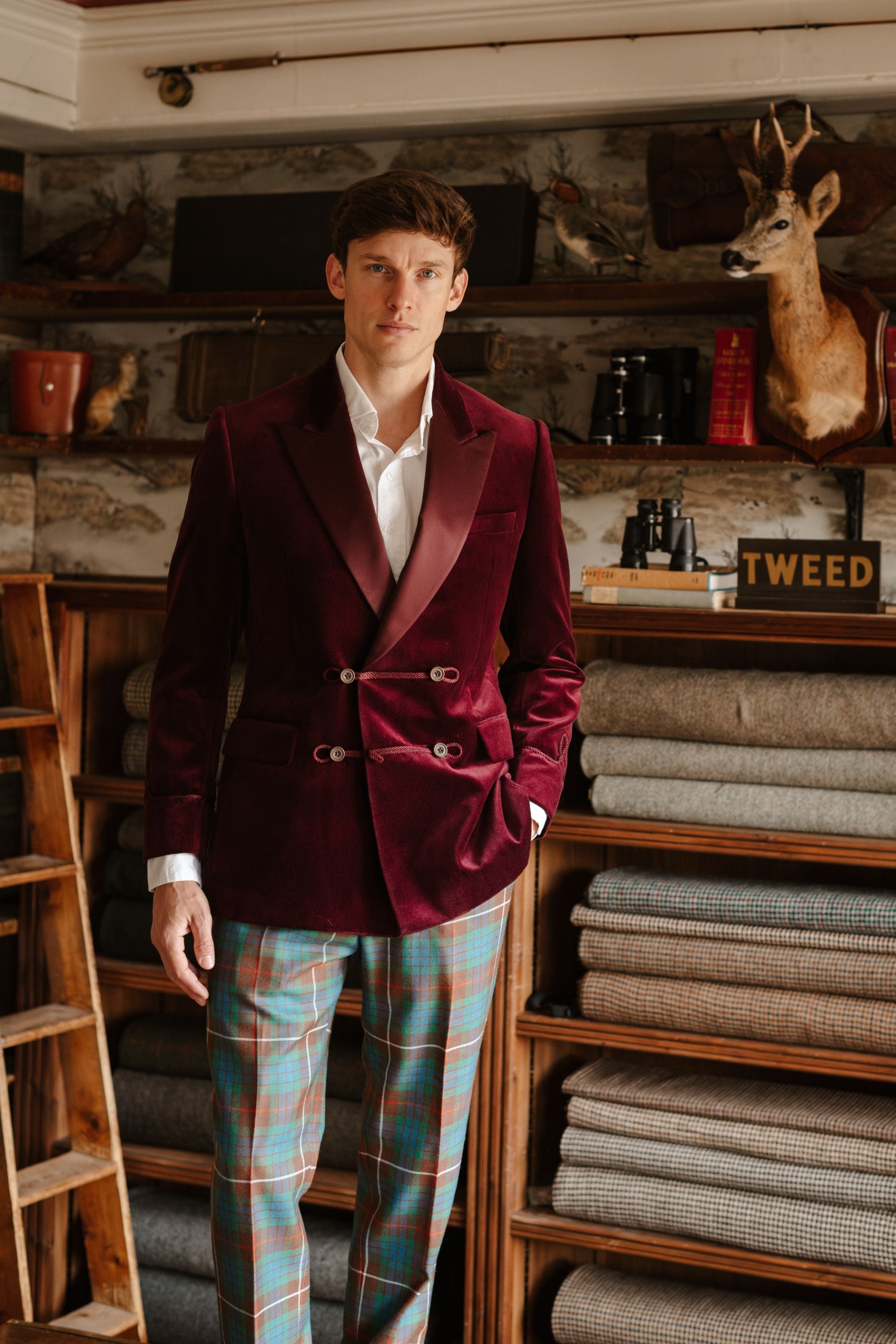 A man wearing a maroon velvet double-breasted blazer, white shirt, and plaid trousers stands in a fabric shop with shelves of folded textiles and rustic decor, including a mounted deer head, behind him.