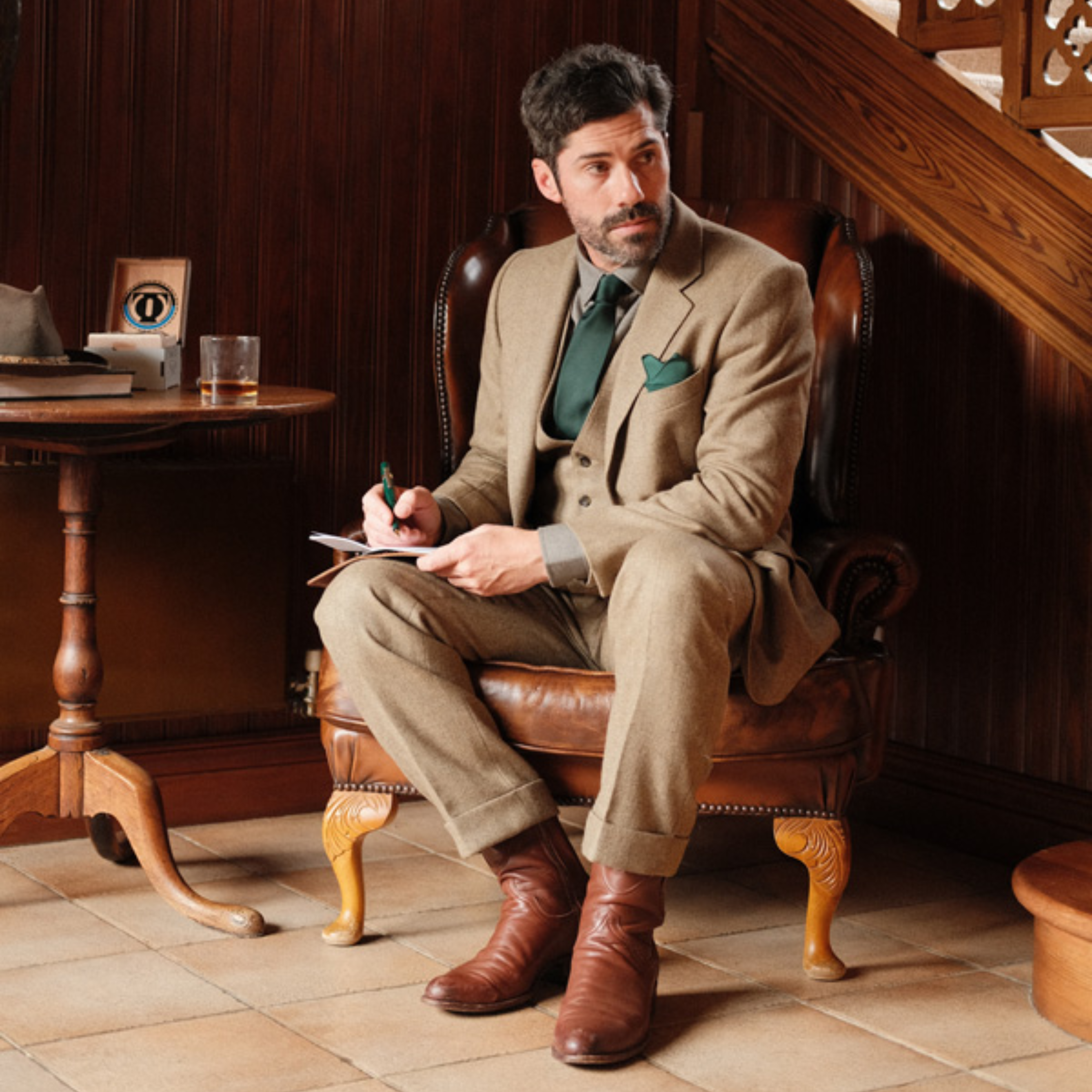 A man with dark hair and a beard, dressed in a tan suit with a green tie and pocket square, sits on a leather chair holding a notepad and pen. A wooden table with a drink and hat is beside him, and stairs are in the background.