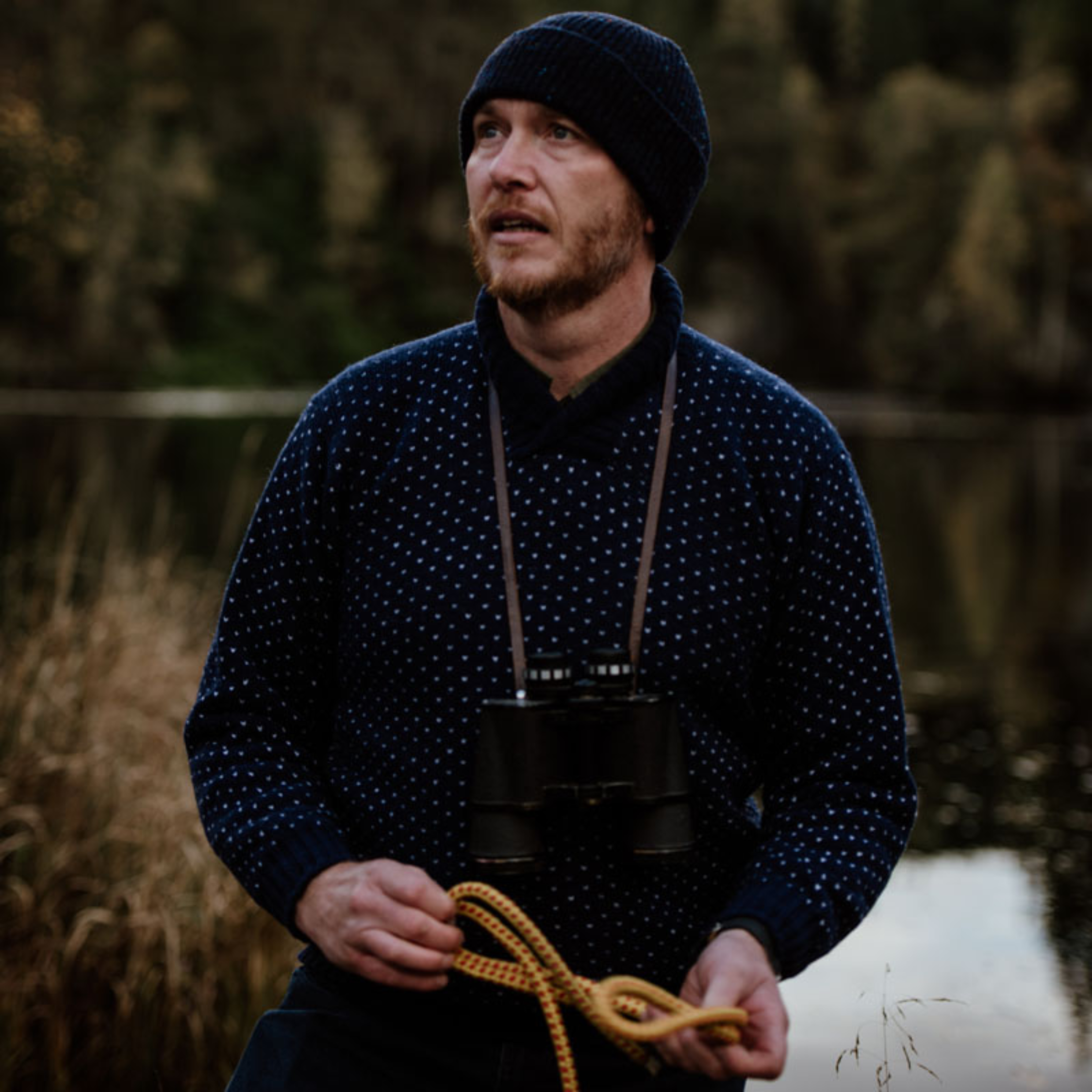 A man wearing a navy beanie and polka-dot sweater stands outdoors near water, holding a yellow rope and binoculars. The background features tall grass and trees.