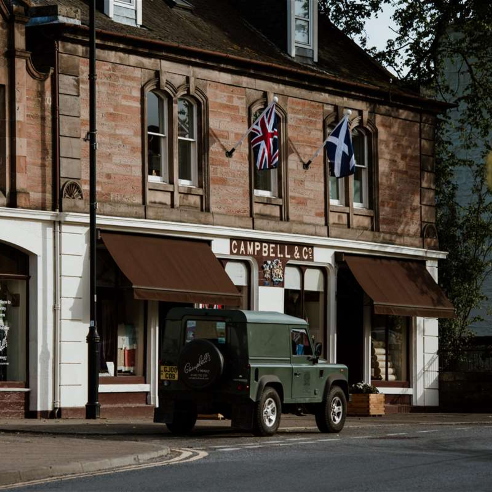 A green Land Rover is parked outside Campbell & Co, a stone building with brown awnings and British and Scottish flags displayed above the entrance.