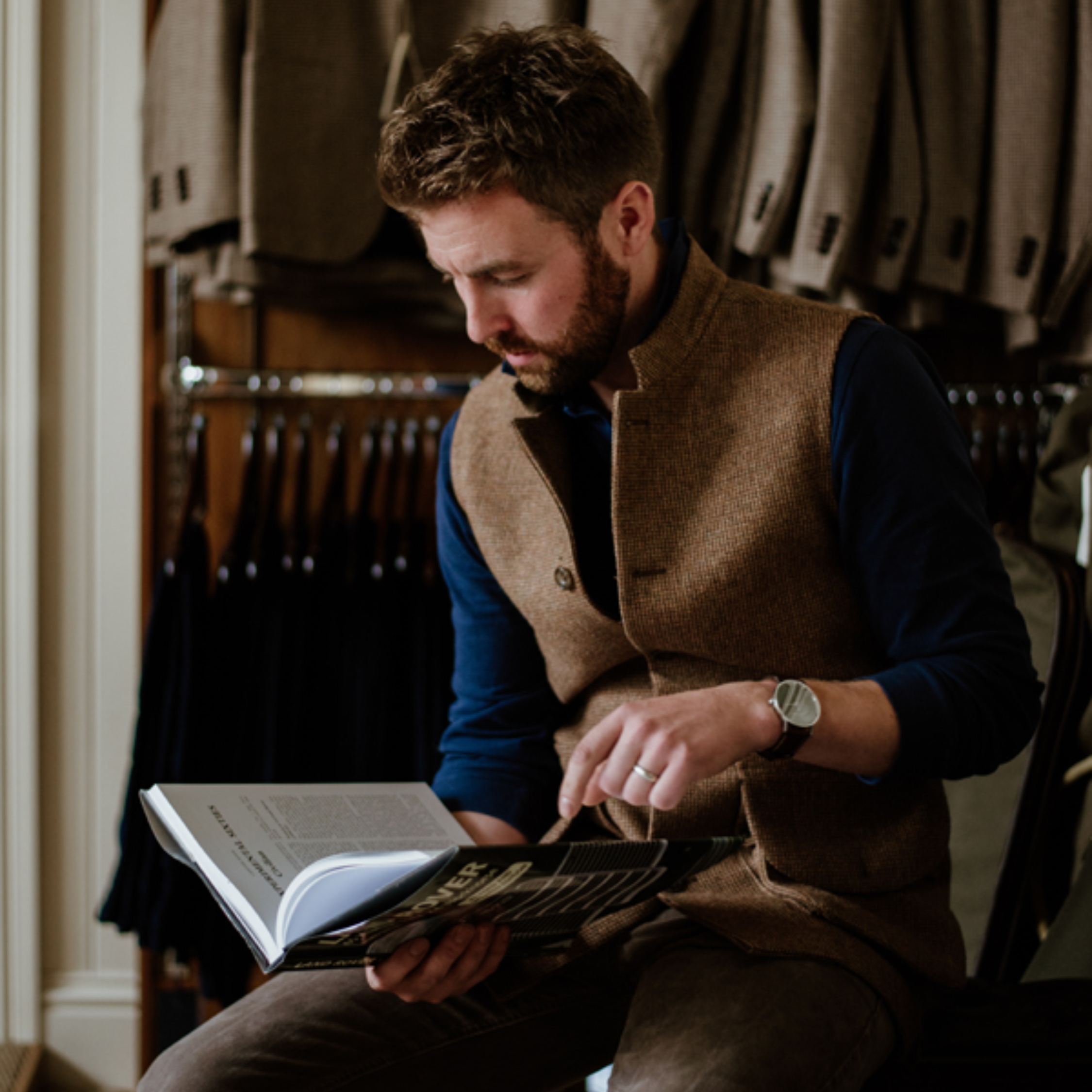 A man with a beard sits indoors, reading an open book. He wears a brown vest over a dark shirt and has a watch on his left wrist. Behind him, beige jackets are hanging on a rail.