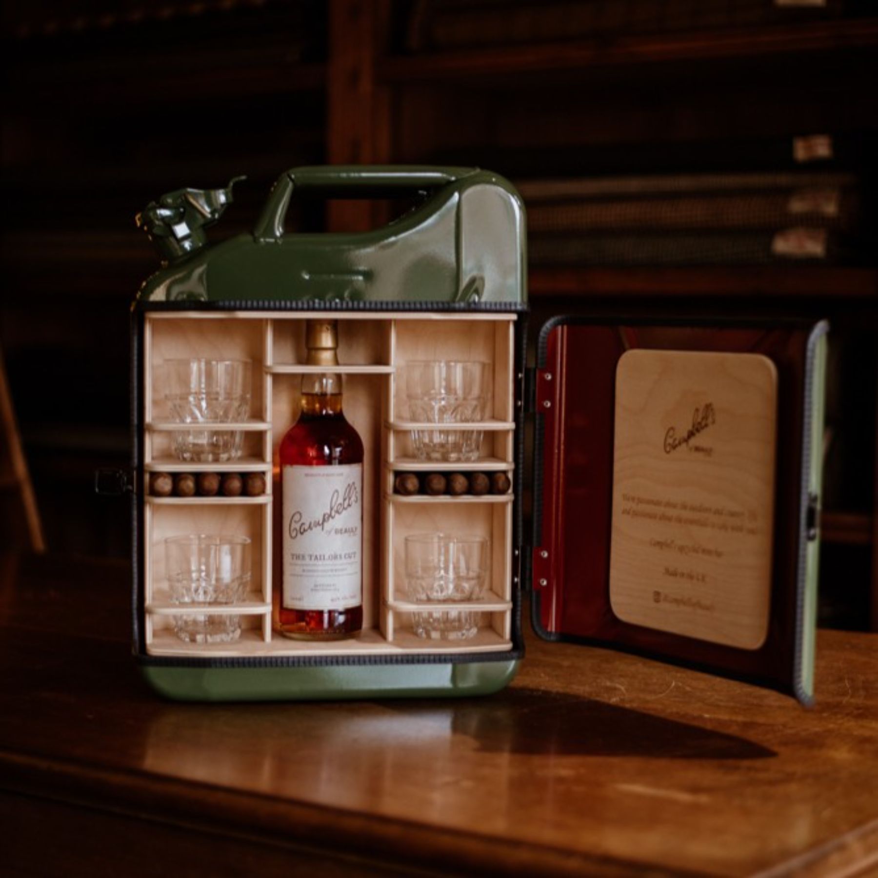 A green metal jerrycan repurposed as a mini bar, holding a bottle of Campbell’s whisky, four glasses, and several whisky stones, with the lid open to display the contents on a wooden table.