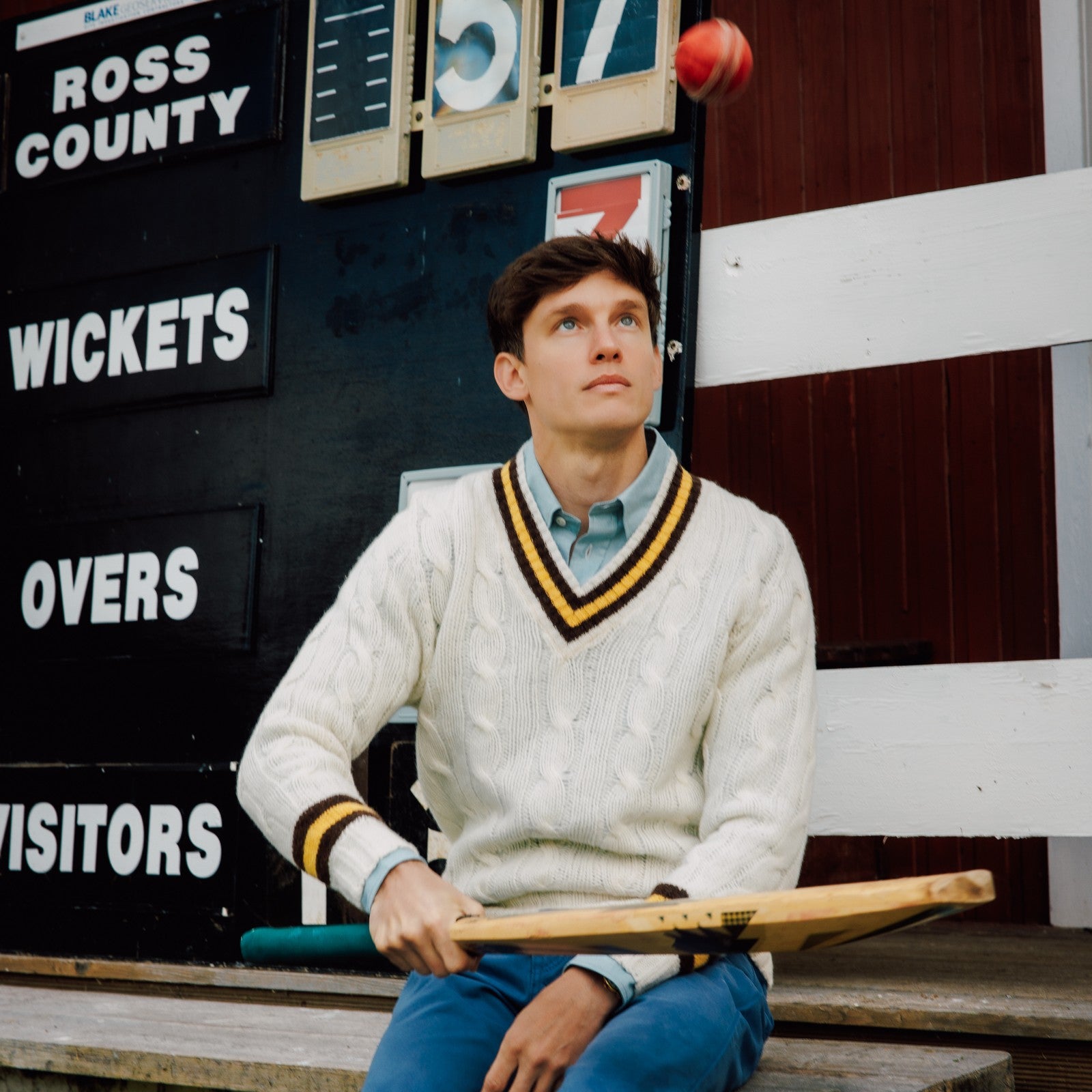 A young man in a cricket sweater sits in front of a scoreboard, tossing a red cricket ball in the air while holding a bat, looking upward with a thoughtful expression.