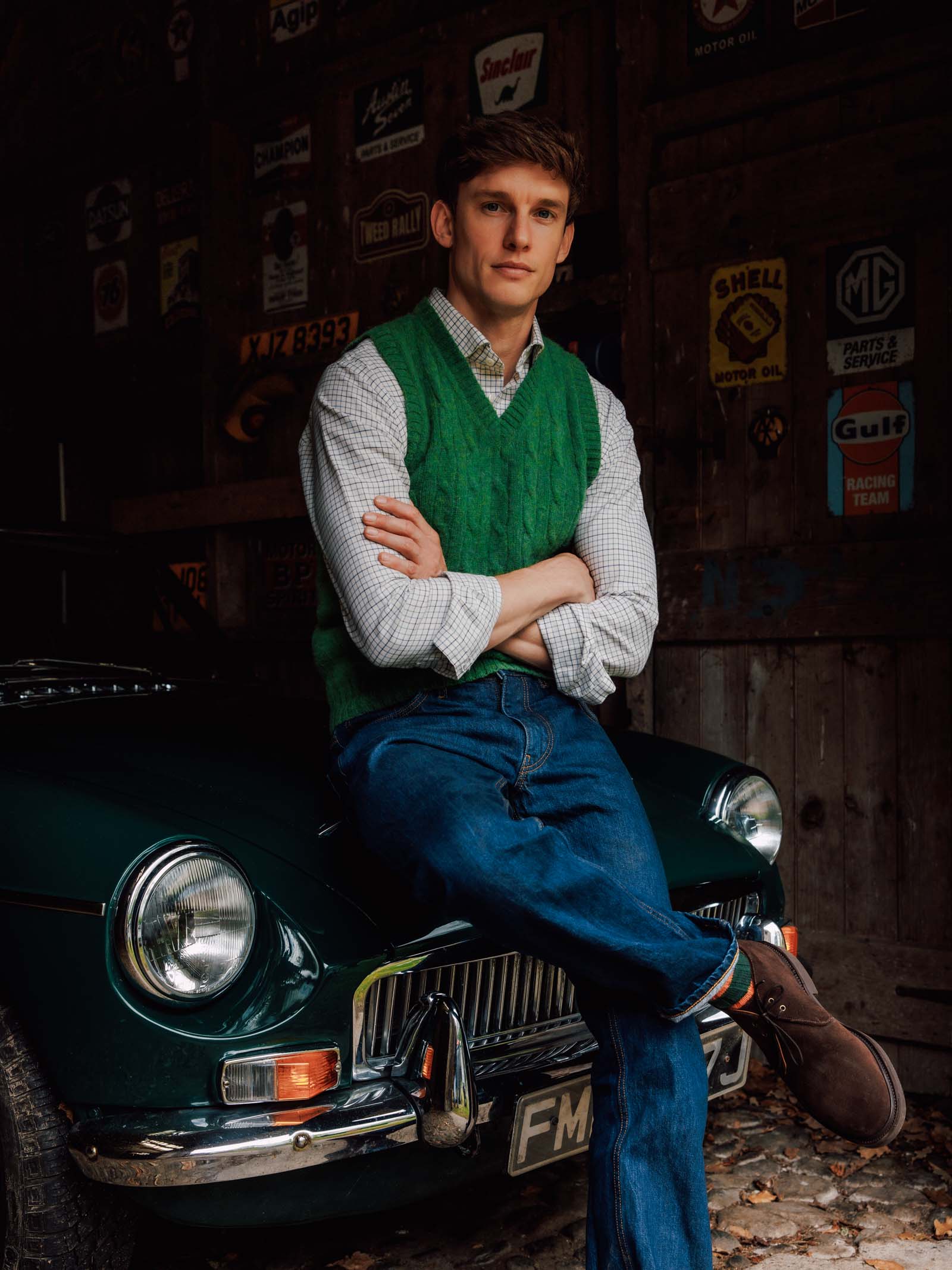 A young man in a green sweater vest and jeans, sporting Campbell’s of Beauly Lomond Boots in chocolate brown suede with sturdy Goodyear welted construction, sits on the hood of a vintage car inside a rustic garage decorated with retro signs.