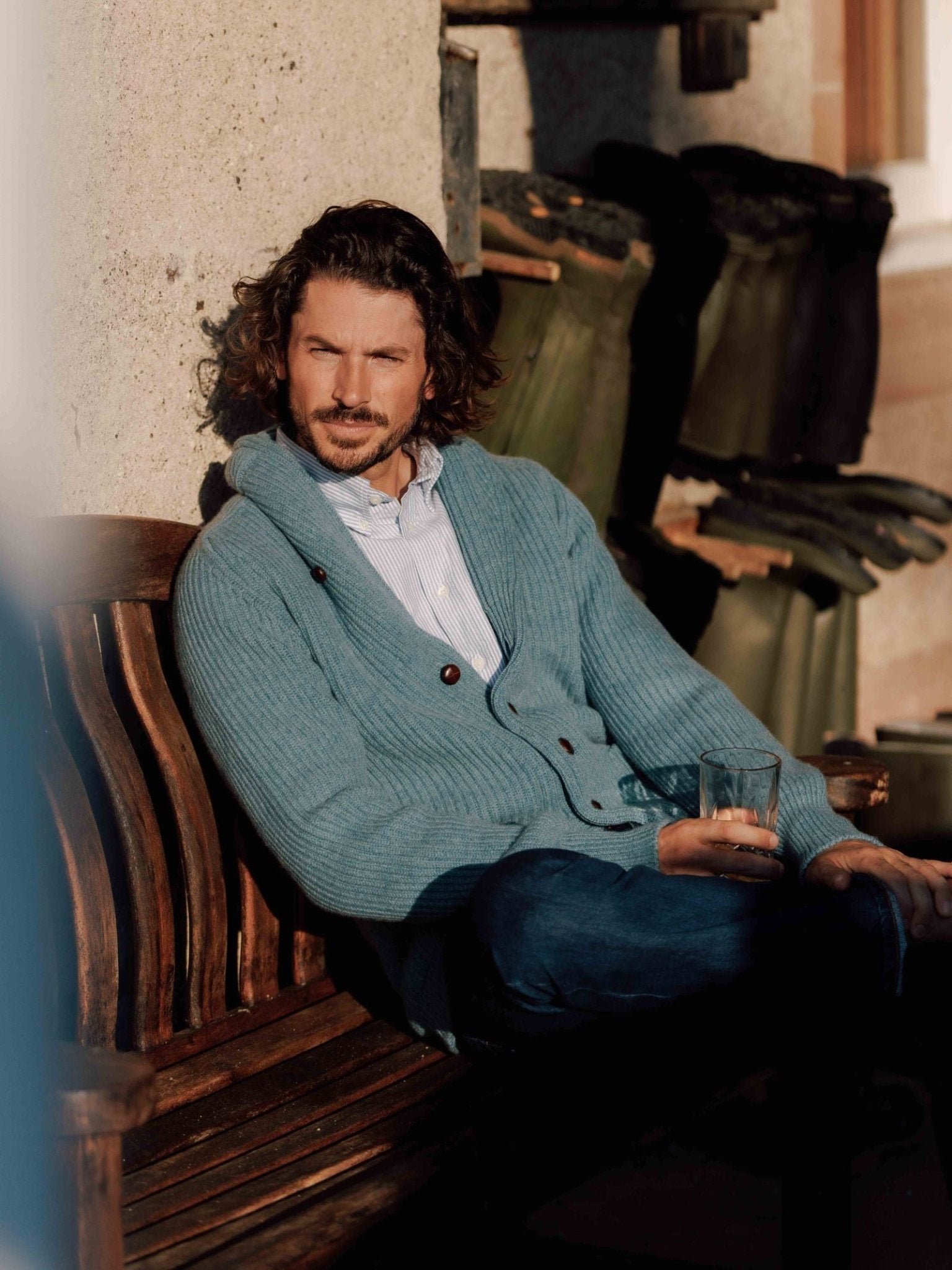 A man with curly hair sits on a wooden bench outdoors, wearing a light blue Campbell's of Beauly Cashmere Shawl Collar Cardigan and jeans, holding a glass and gazing thoughtfully into the distance.