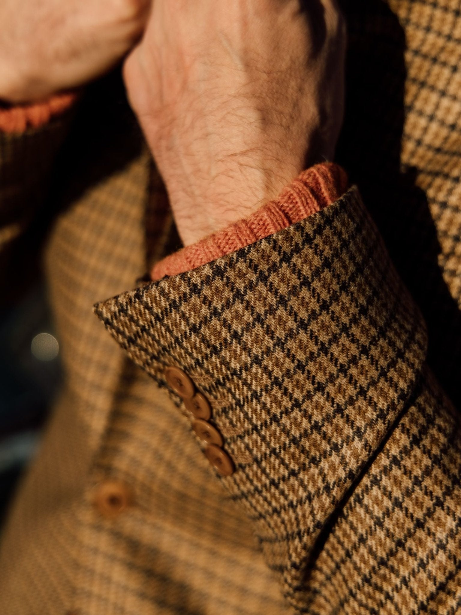 A close-up of a hand adjusting the cuff of the Campbell's of Beauly Auld Stock Gun Club Jacket in brown and beige, with a rust-colored knit sweater visible underneath.