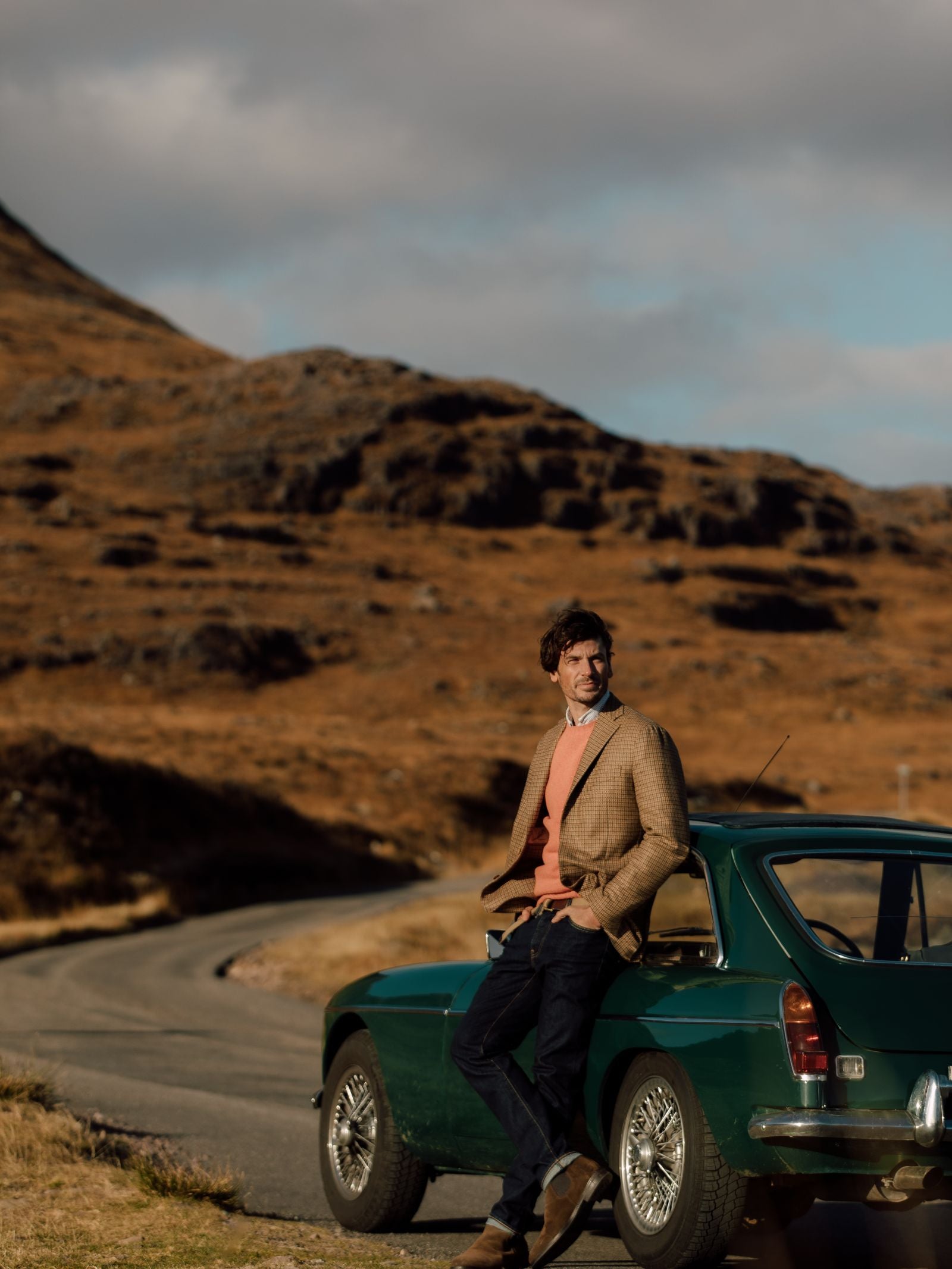 A man in a Campbell's of Beauly Auld Stock Gun Club Jacket leans against a vintage green car on a winding road, backed by rocky brown hills beneath a partly cloudy sky.