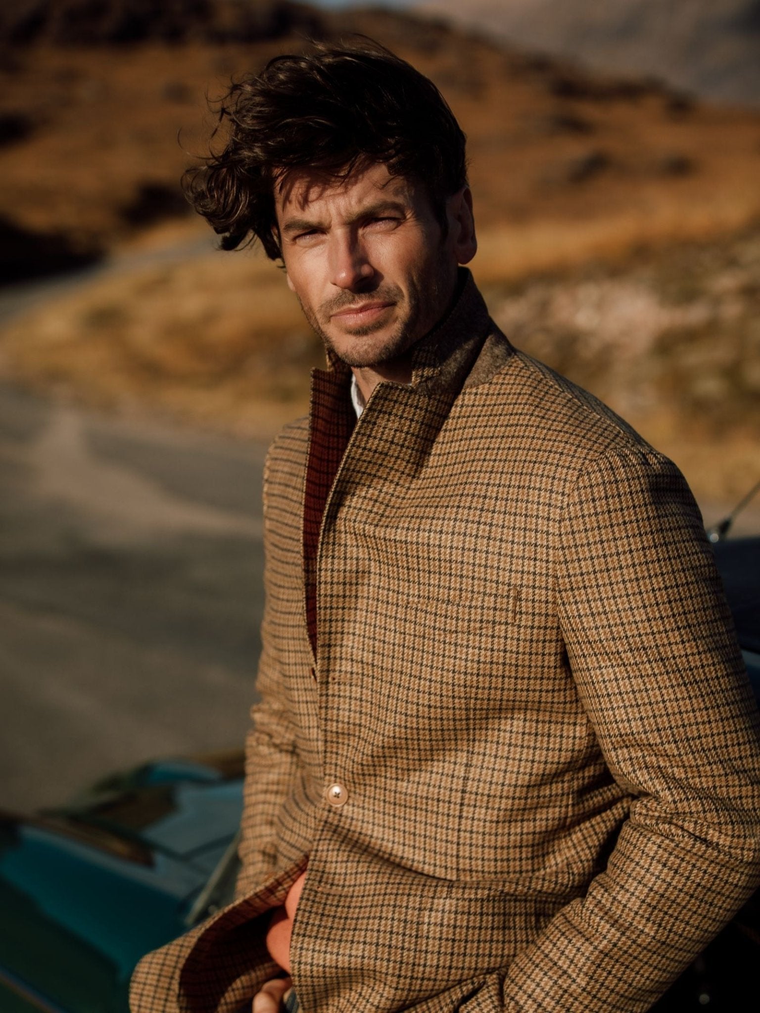 A man with tousled hair sits outdoors in sunlight, wearing the Auld Stock Gun Club Jacket by Campbell's of Beauly, framed by a blurred landscape of hills and dry grass in the background.