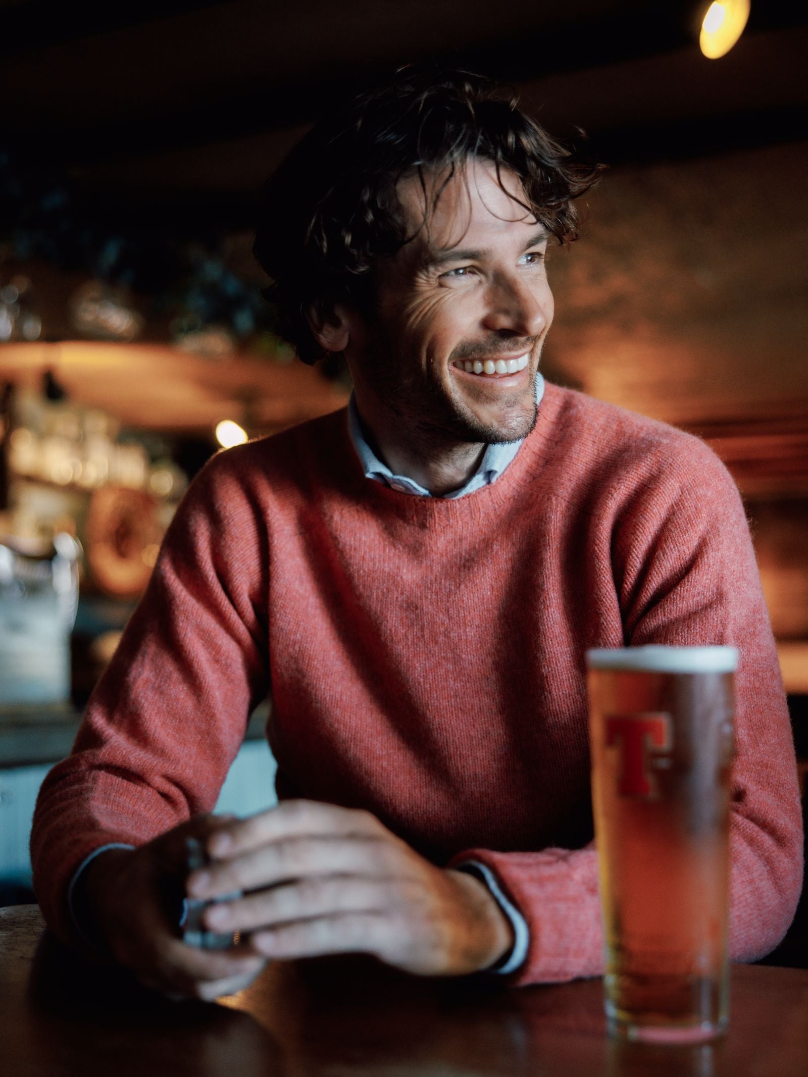 A smiling man in a Campbell's of Beauly Shetland Jumper sits at a wooden table in a cozy bar, holding something. In front of him is a tall glass of beer with a red T logo. The background is softly blurred.