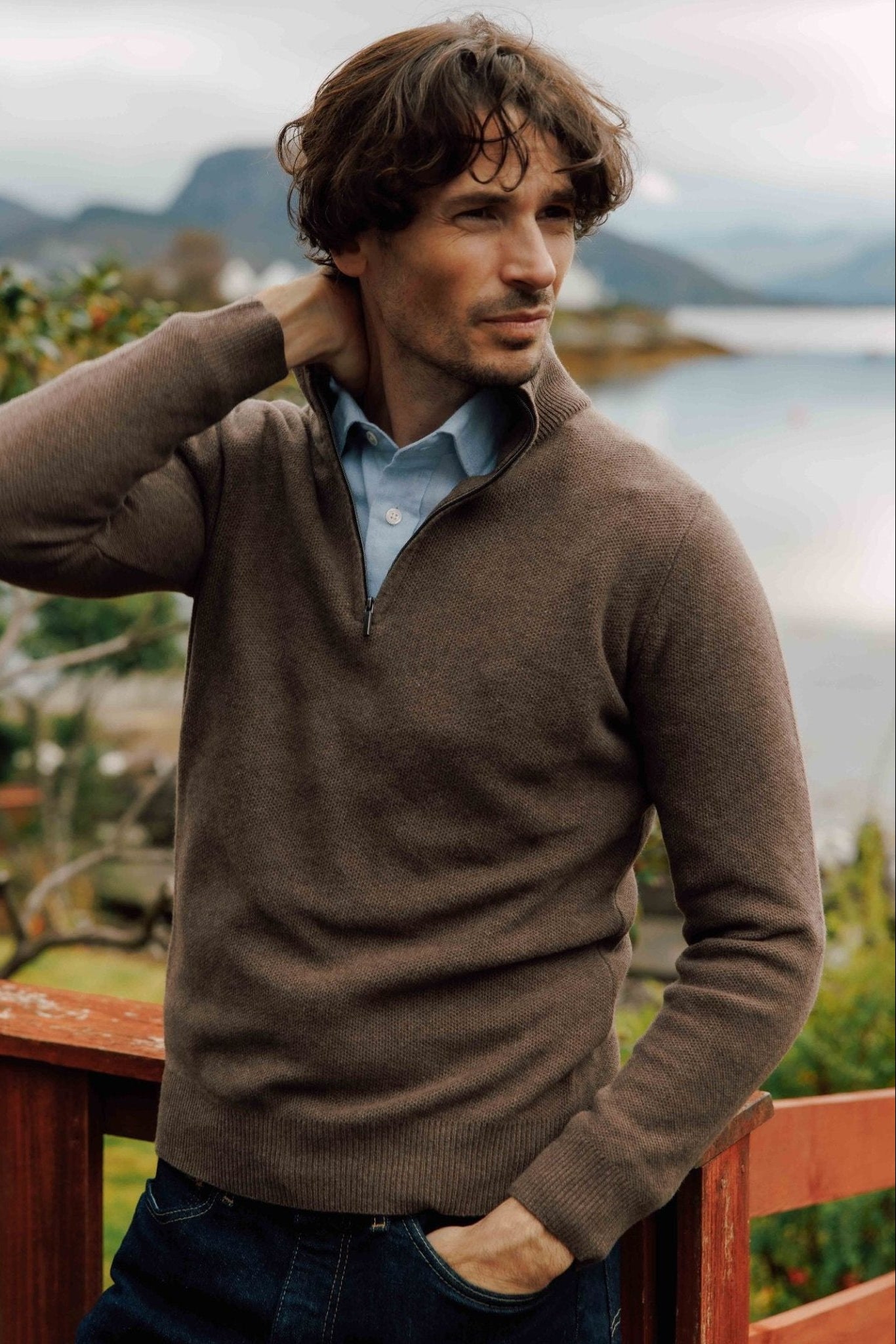 A man with wavy brown hair, wearing the Campbell's of Beauly Auld Stock Piquet Quarter Zip Jumper over a light blue shirt, stands by a wooden railing with a scenic lake and mountain landscape in the background.