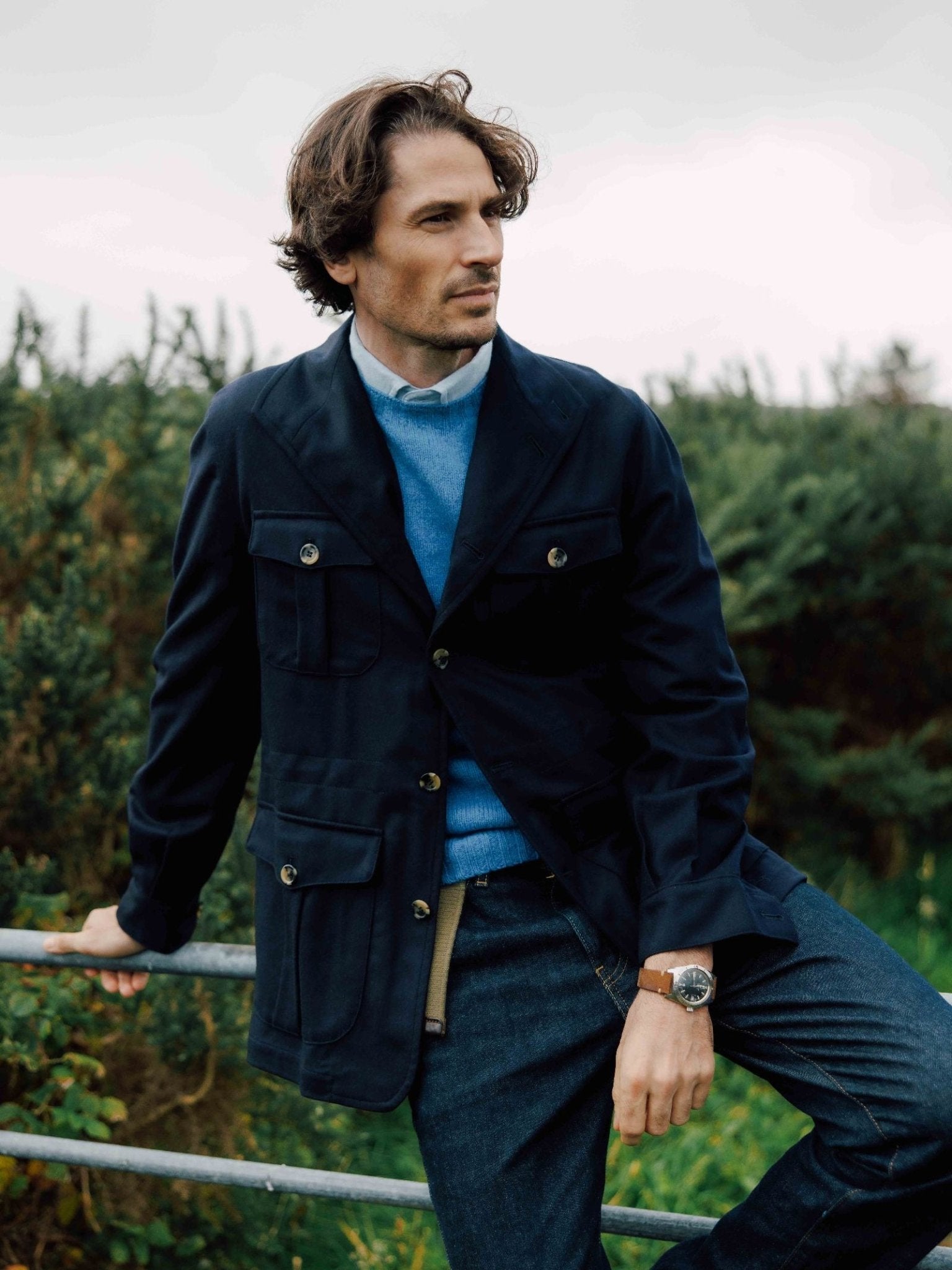 A man with wavy brown hair wears the Auld Stock Field Teba Jacket by Campbell's of Beauly, paired with a blue sweater and denim jeans. He leans on a railing outdoors amid lush greenery under cloudy skies.