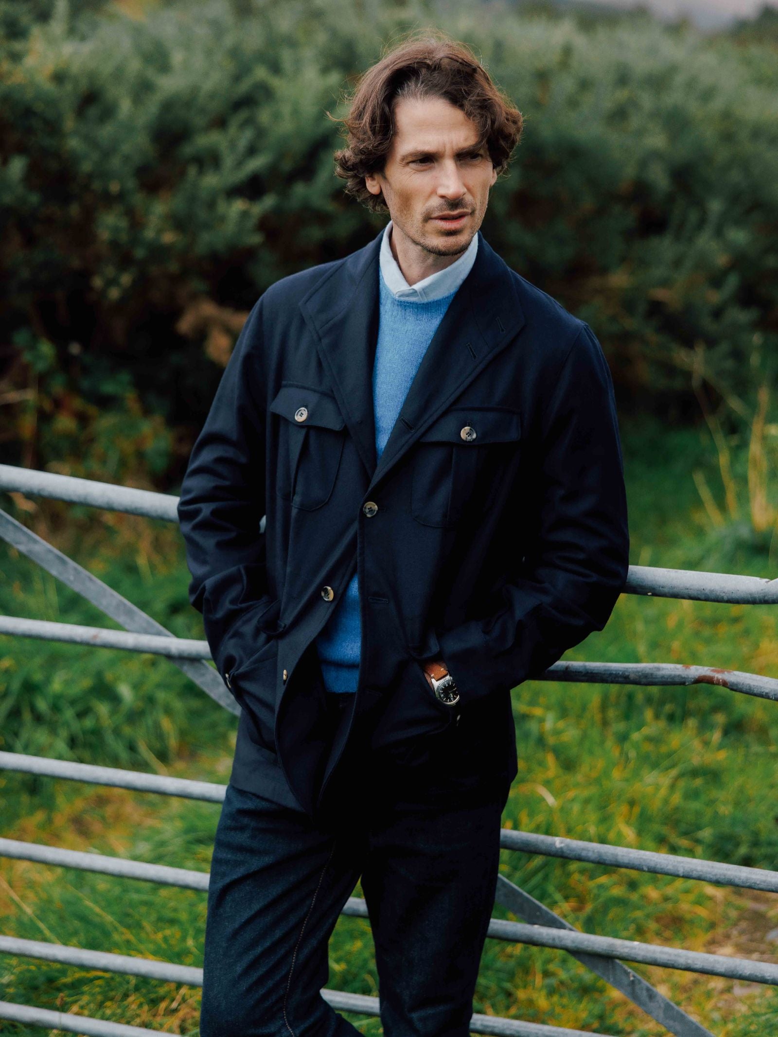 A man with wavy brown hair stands outdoors by a metal gate, wearing the Campbell's of Beauly Auld Stock Field Teba Jacket over a blue sweater and collared shirt, hands in his pockets amid lush greenery.