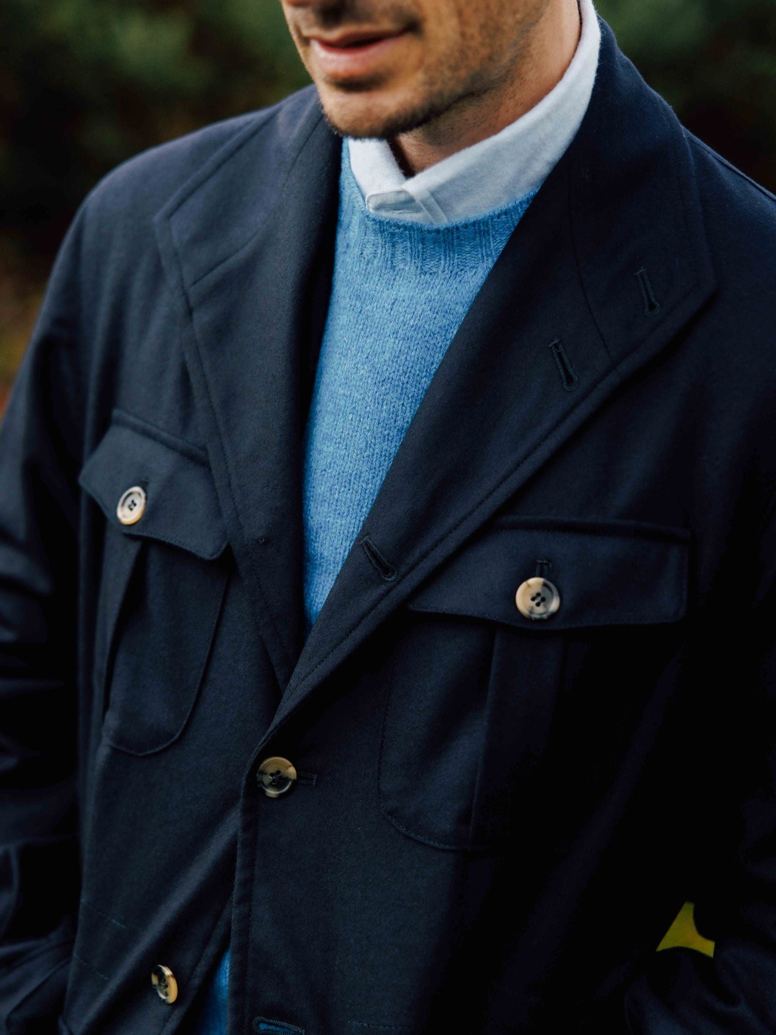 A man wearing the Campbell's of Beauly Auld Stock Field Teba Jacket stands outdoors over a light blue sweater and white shirt, with blurred greenery in the background.