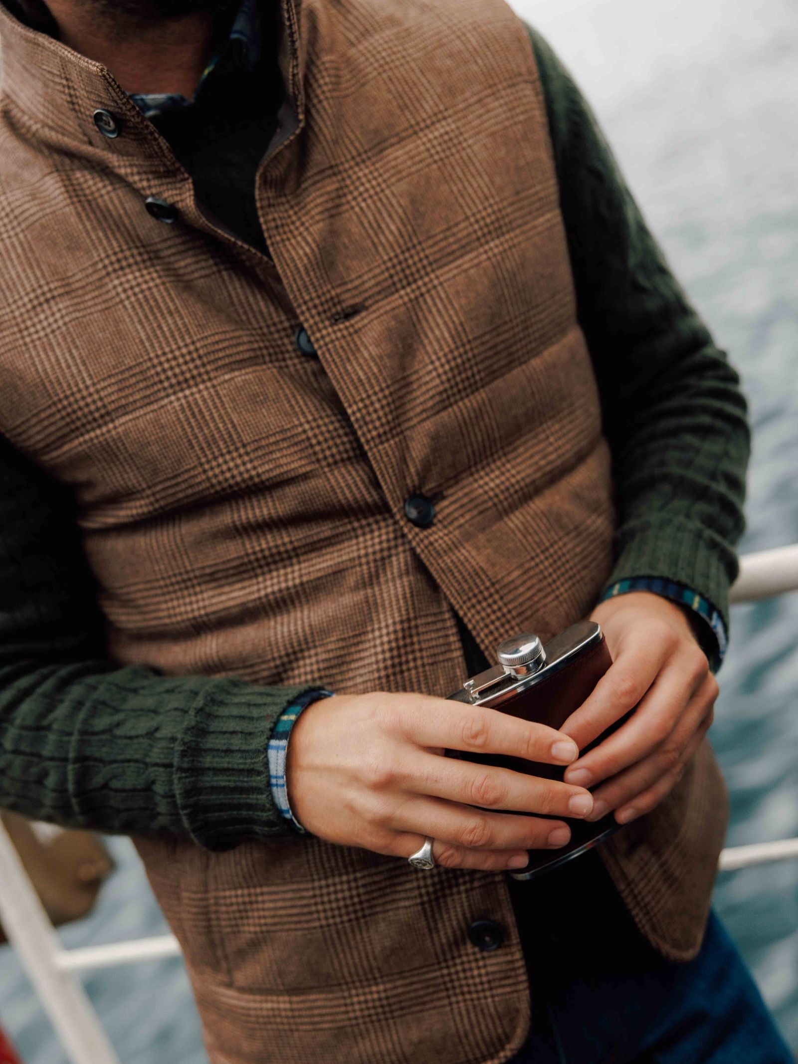 A person in a British tweed look—a Cashmere Gun Club Quilted Gilet by Campbell's of Beauly over a green sweater—stands by the water, holding a metal flask with both hands. The person’s face remains unseen.