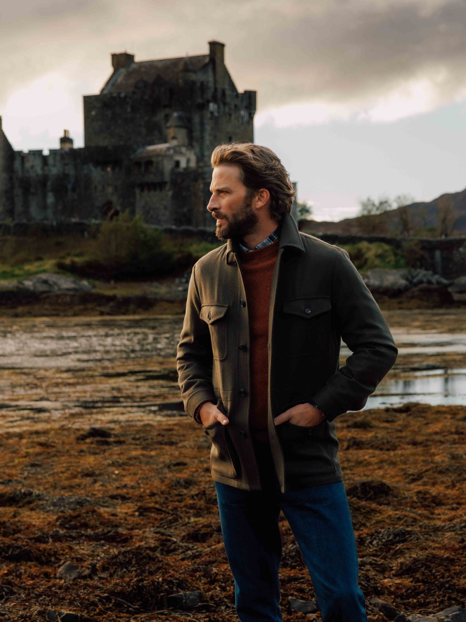 A man with brown hair and a beard stands outdoors in blue jeans and the Auld Stock Winter Aigas Field Jacket by Campbell's of Beauly, with an old stone castle and cloudy sky behind him. He looks to the side, hands in his jacket pockets.