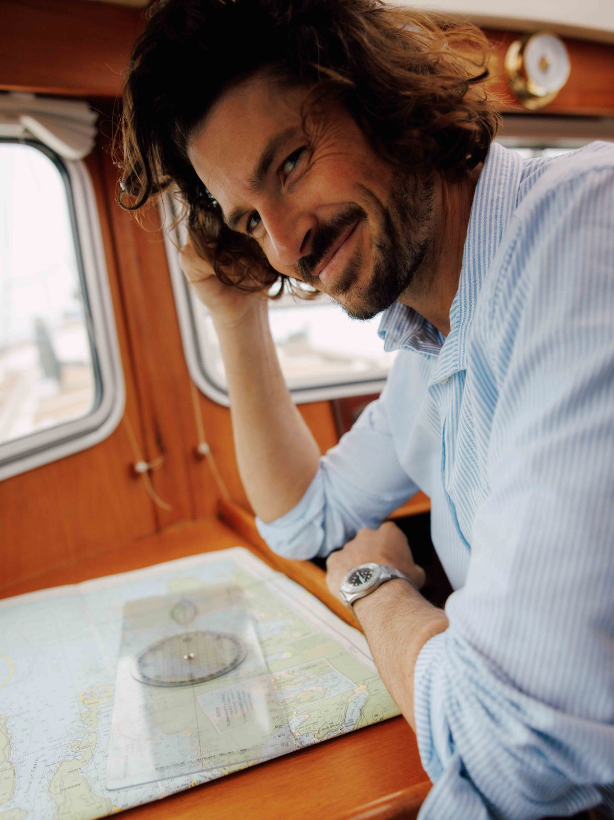 A man with wavy brown hair and a beard wears a Campbells of Beauly Twill Shirt and a watch, smiling as he sits at a wooden table inside a boat with a navigation map and plotting tool.