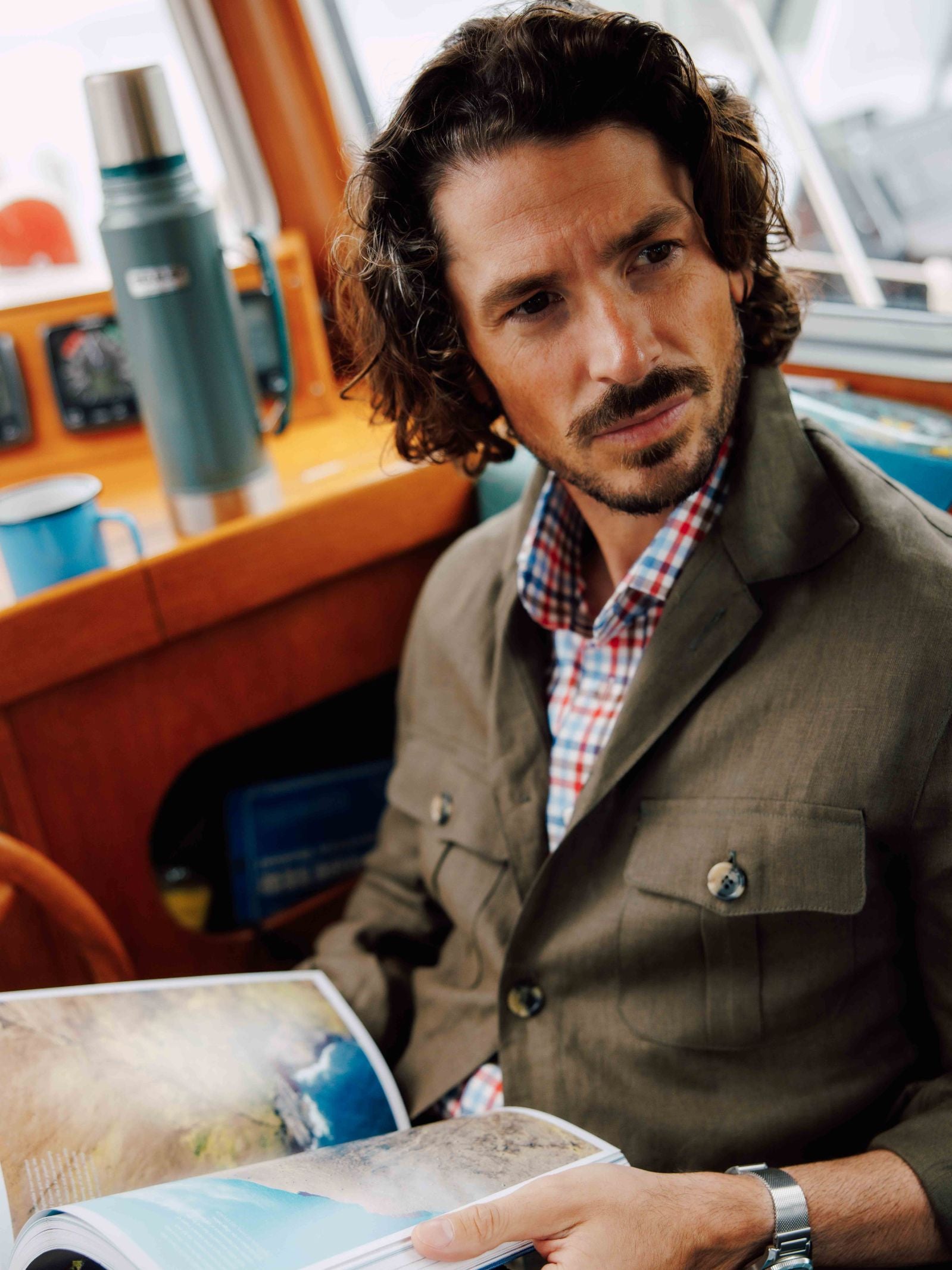 A man with curly hair and a beard, dressed in a plaid shirt and Campbell's of Beauly Linen Safari Jacket, sits indoors holding an open magazine, with a mug, thermos, and navigation equipment in the background.