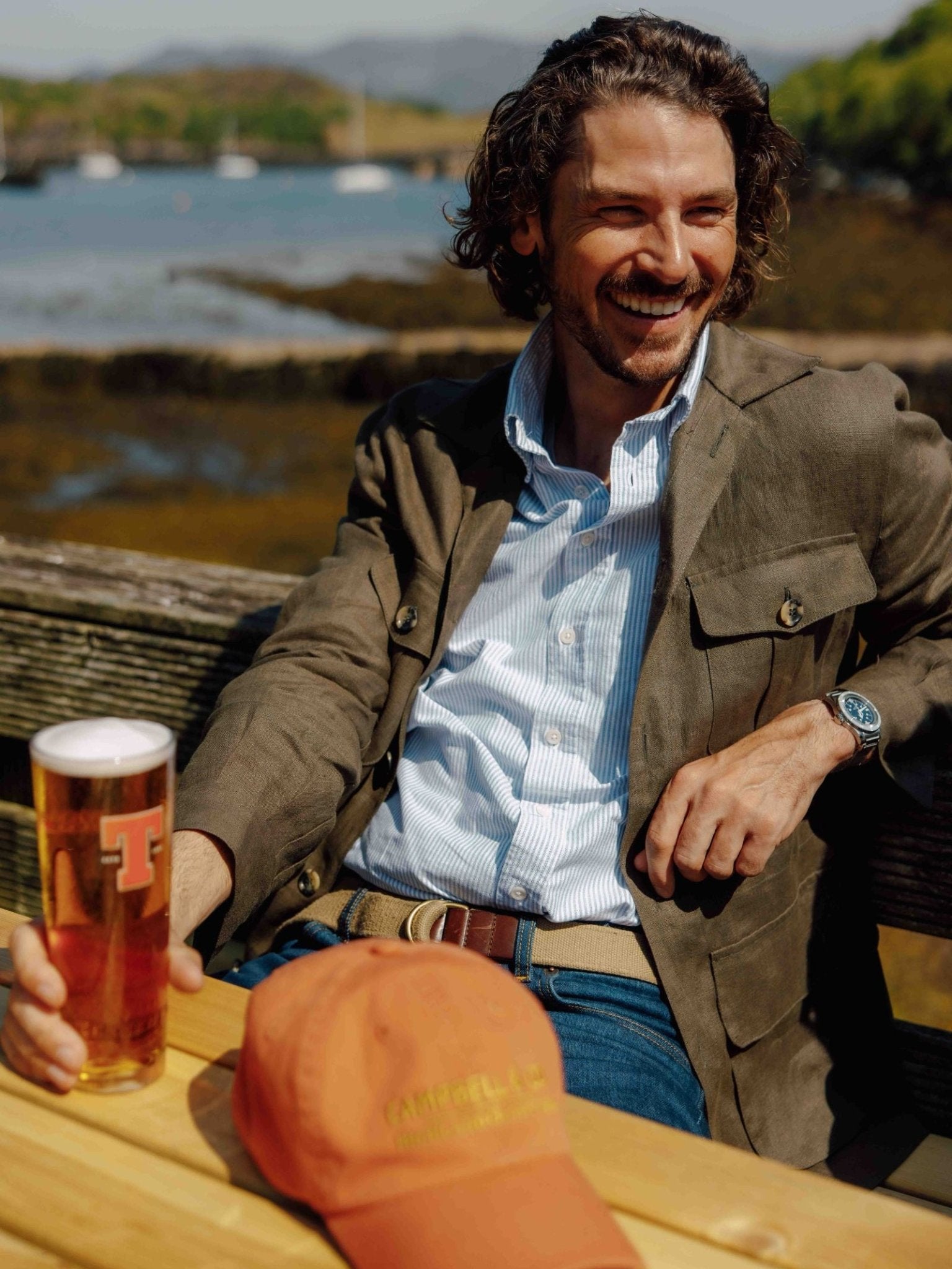 A smiling man with wavy brown hair sits outdoors at a wooden table, holding a pint of beer. He wears jeans, a green blazer, and the Campbell's of Beauly Auld Stock Oxford Shirt. An orange cap rests on the table; water and hills are in the background.