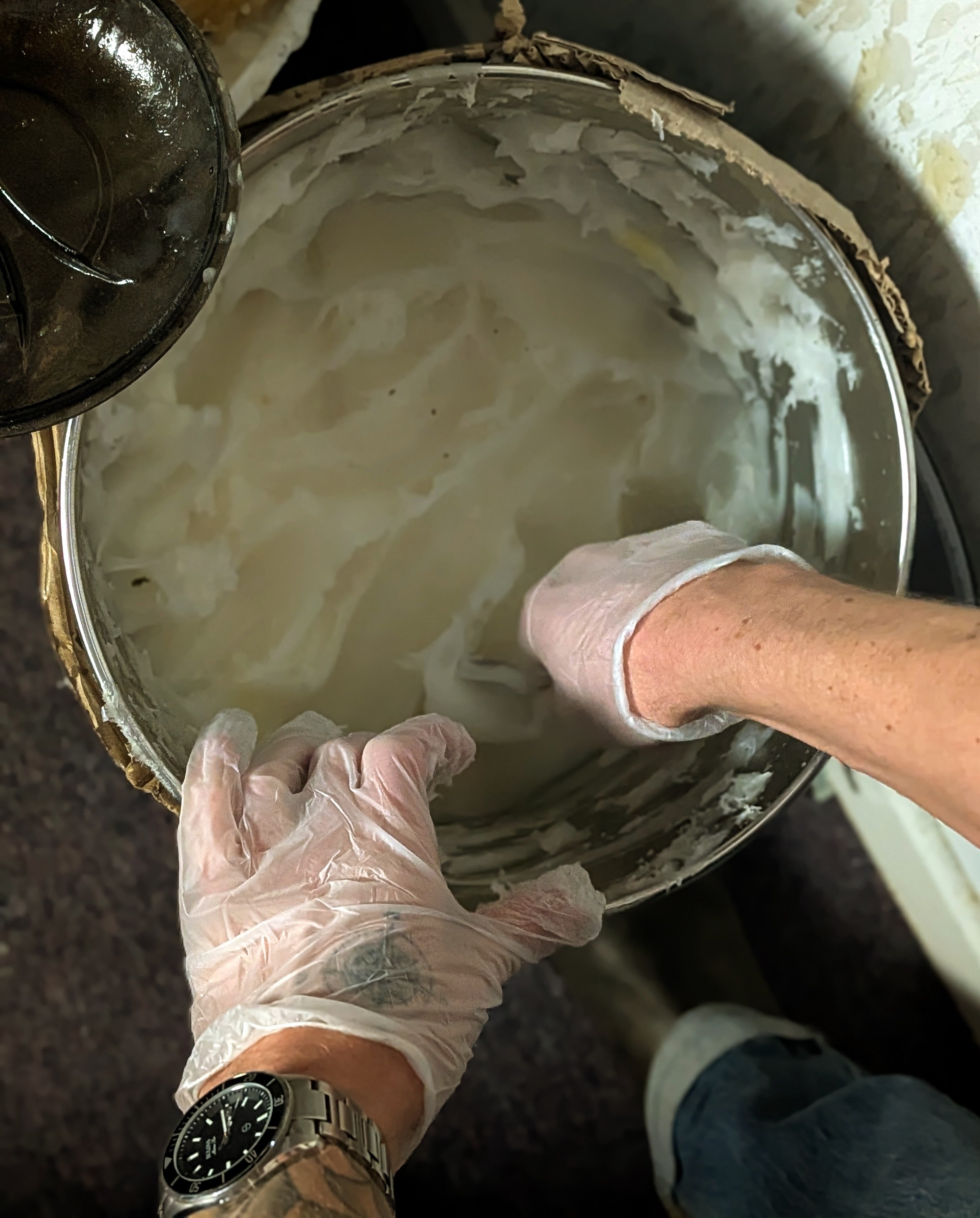 A person wearing disposable gloves and a watch is scooping or mixing a thick, white substance in a large, open container. The persons hands are visible, one inside the container and the other holding its edge.