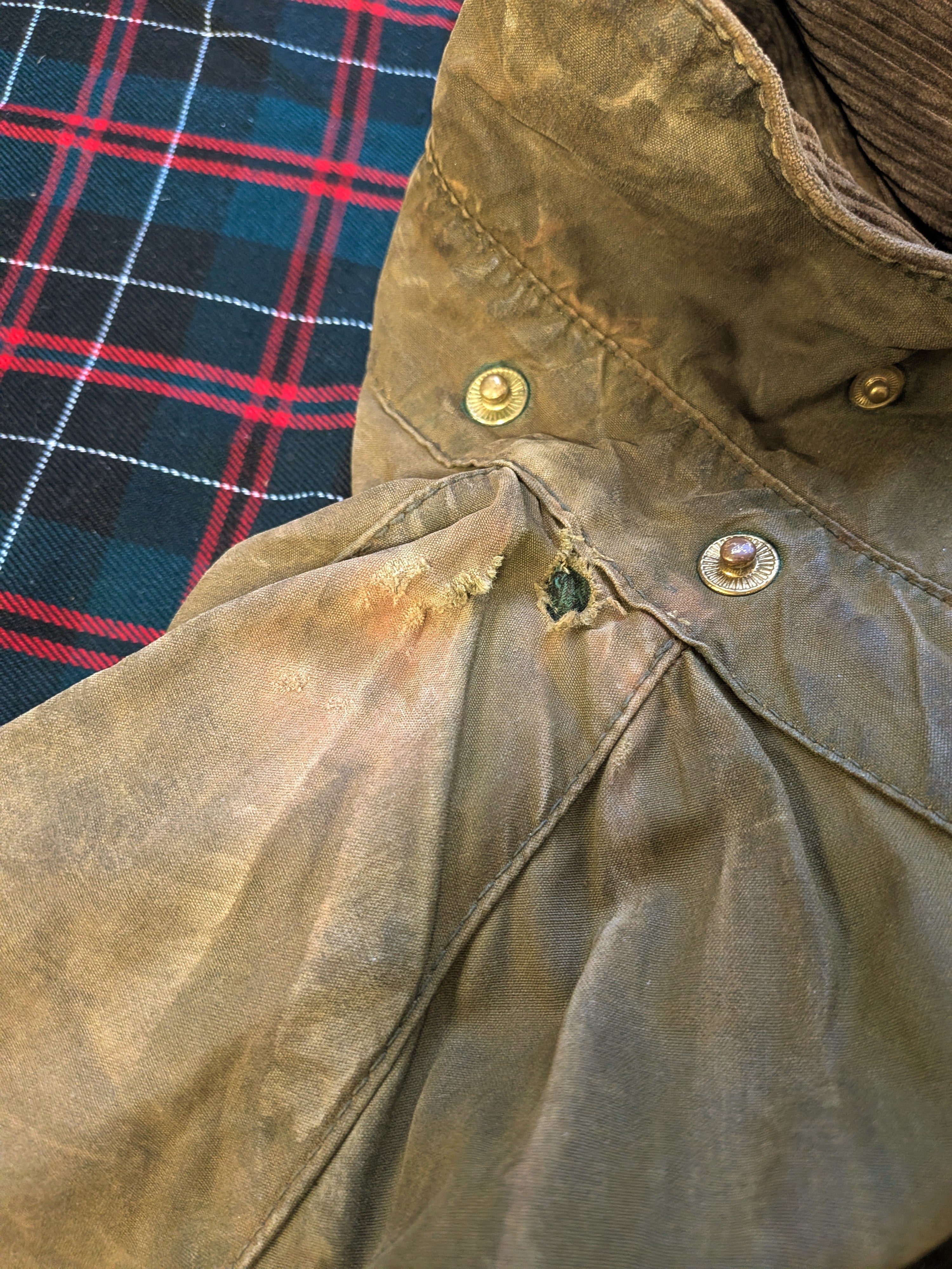 A close-up of a worn brown jacket with a tear and yellowish stains near the sleeve seam, lying on a red and blue plaid fabric.
